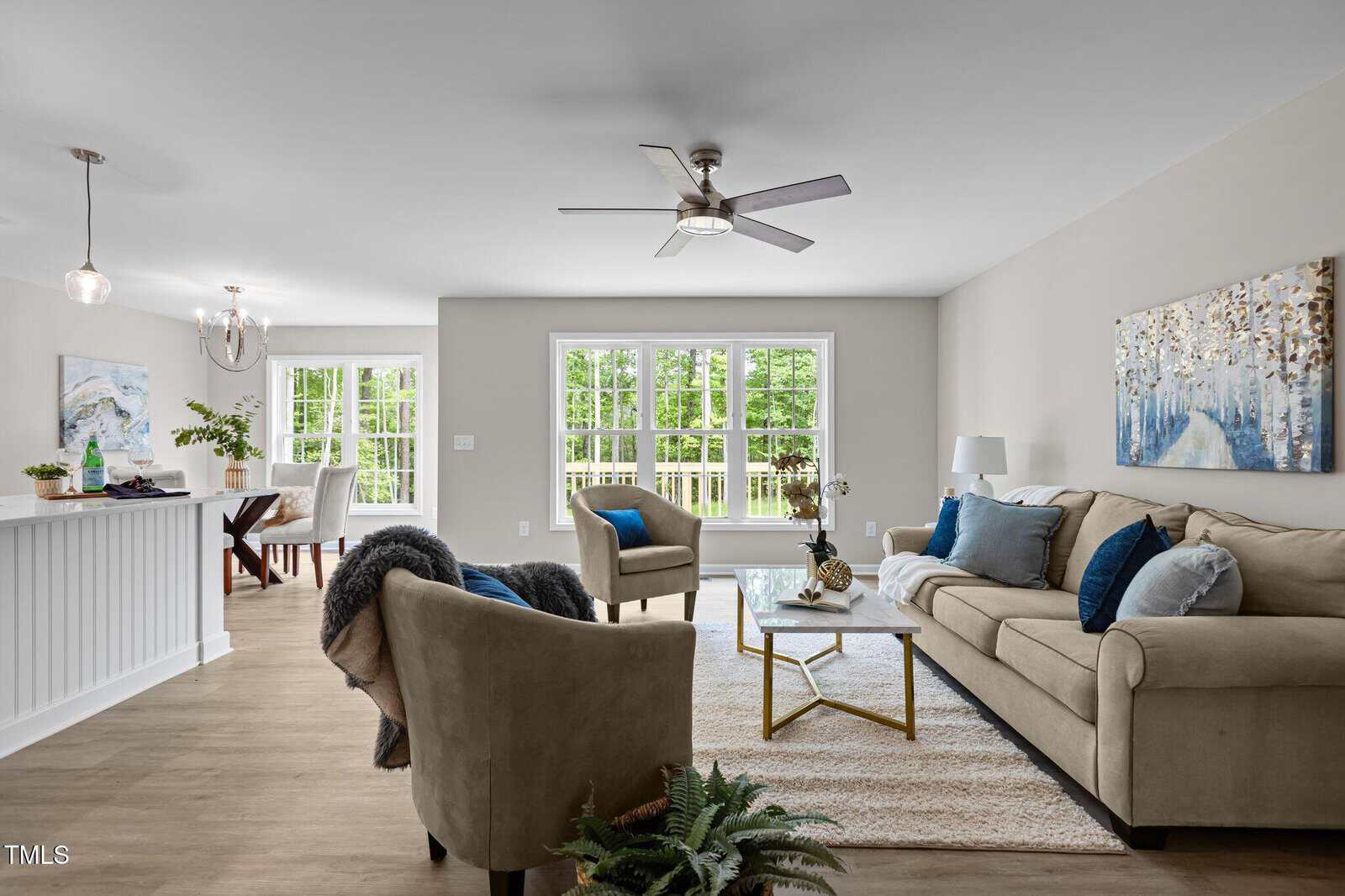 Modern living room with beige sofa, blue pillows, armchairs, coffee table, area rug, and artwork on the wall. Dining area and large windows with a view of greenery in the background. Neutral tones and natural light.