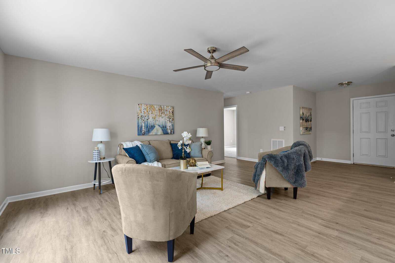 A modern living room with light wood floors, beige walls, a ceiling fan, a sofa, two armchairs, a coffee table, lamps, and wall art above the sofa. A doorway and white front door are visible in the background.