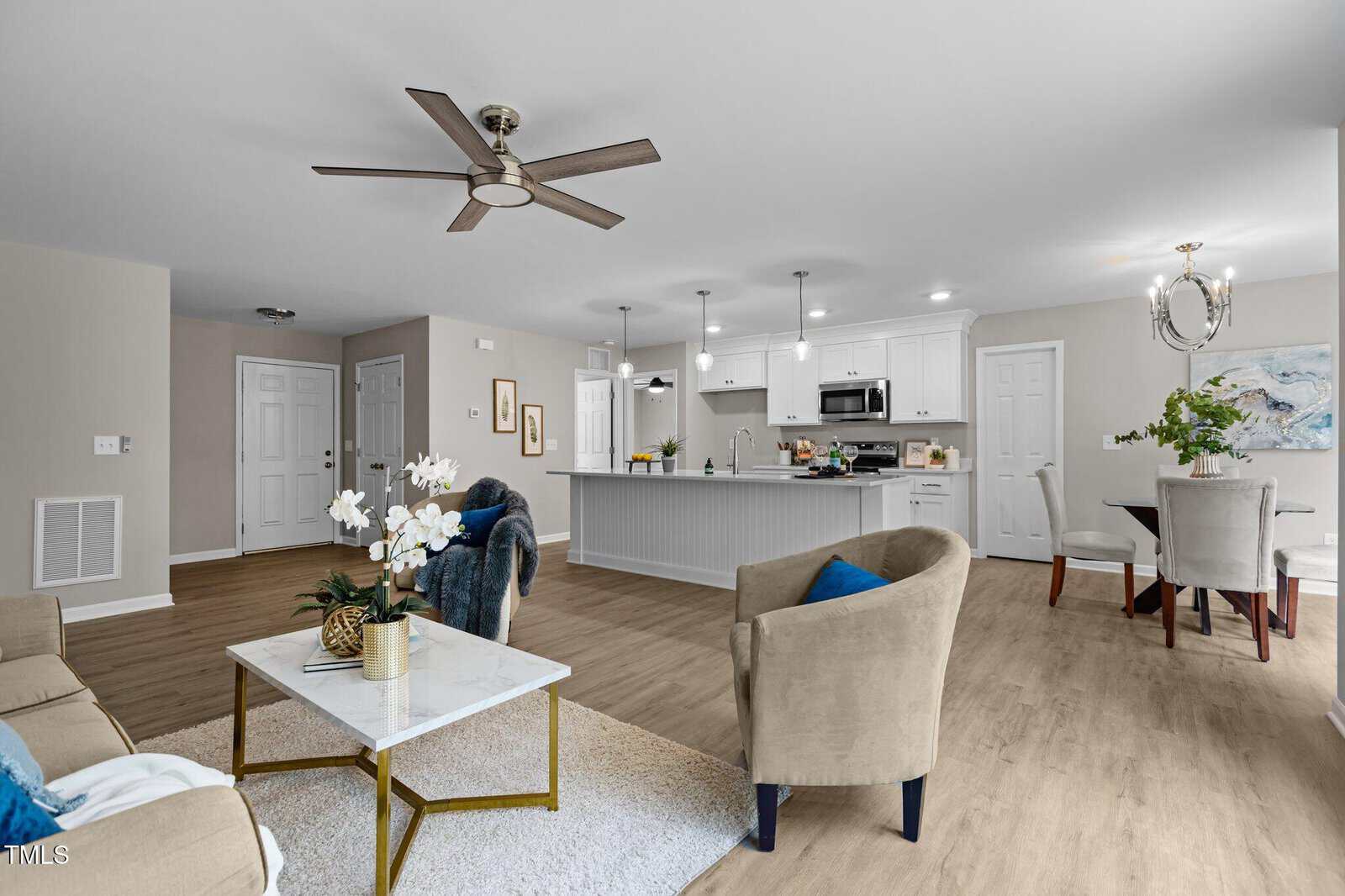 Modern open-concept living space with beige walls, wood flooring, a cozy sitting area, kitchen with white cabinets, island, stainless steel appliances, and a dining area with a round table and chandelier.