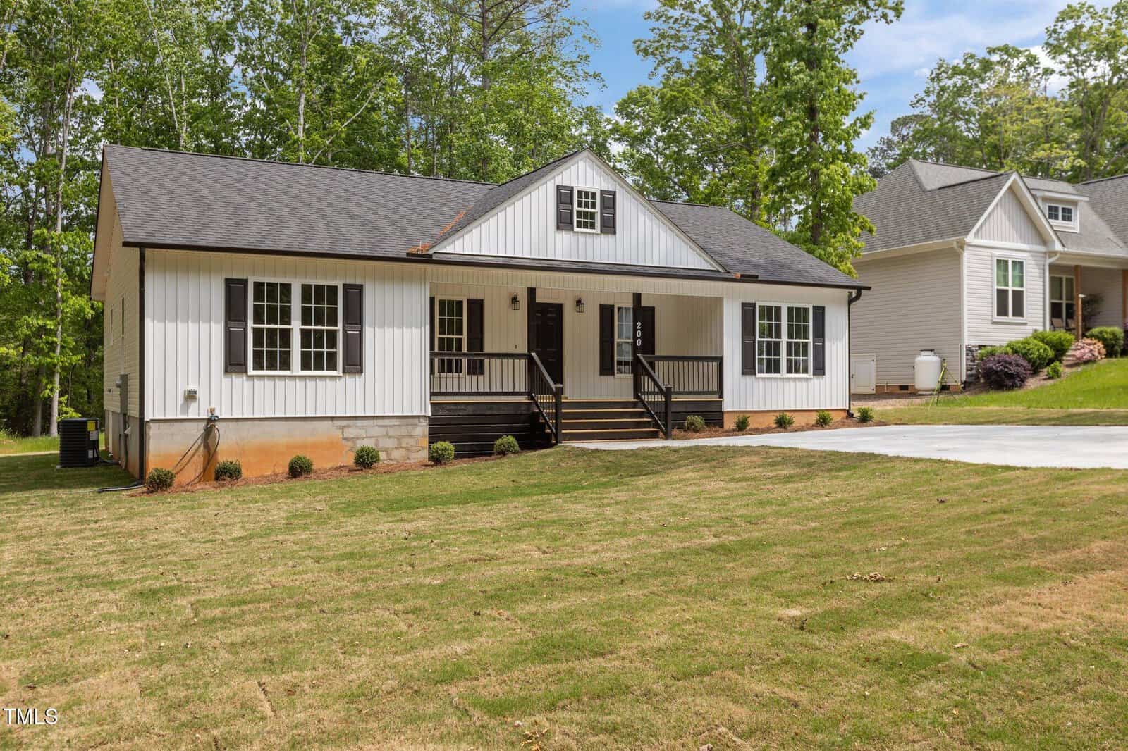 Single-story white house with black trim, a gable roof, covered front porch with black railings, and wide driveway. The front yard has patchy grass, small shrubs, and trees in the background.