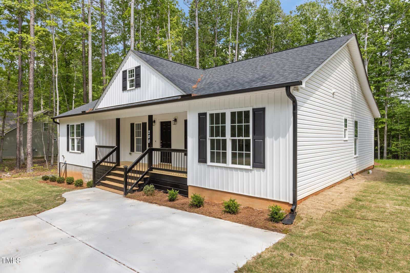 A single-story white house with black shutters and a gable roof sits on a grassy lot. The front porch has black railings and steps leading to a concrete driveway. Tall trees surround the property.
