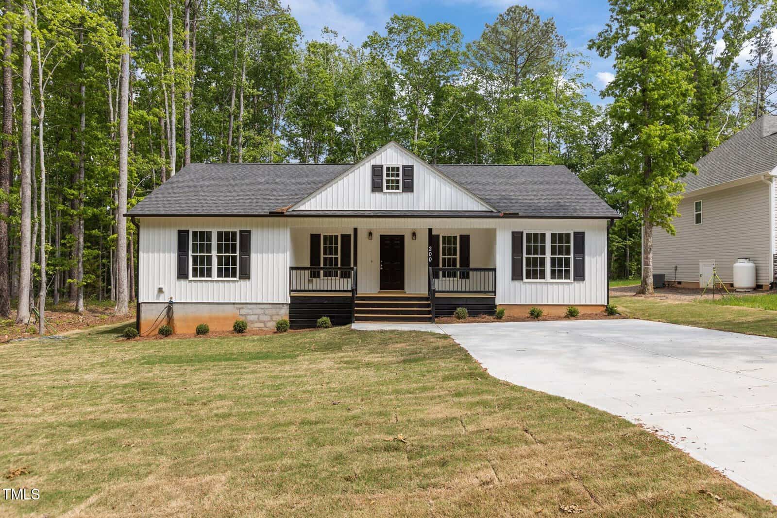 A single-story house with vertical white siding, black trim, and a covered front porch with stairs. It is surrounded by a mowed lawn, trees, and a concrete driveway leading to the entrance.