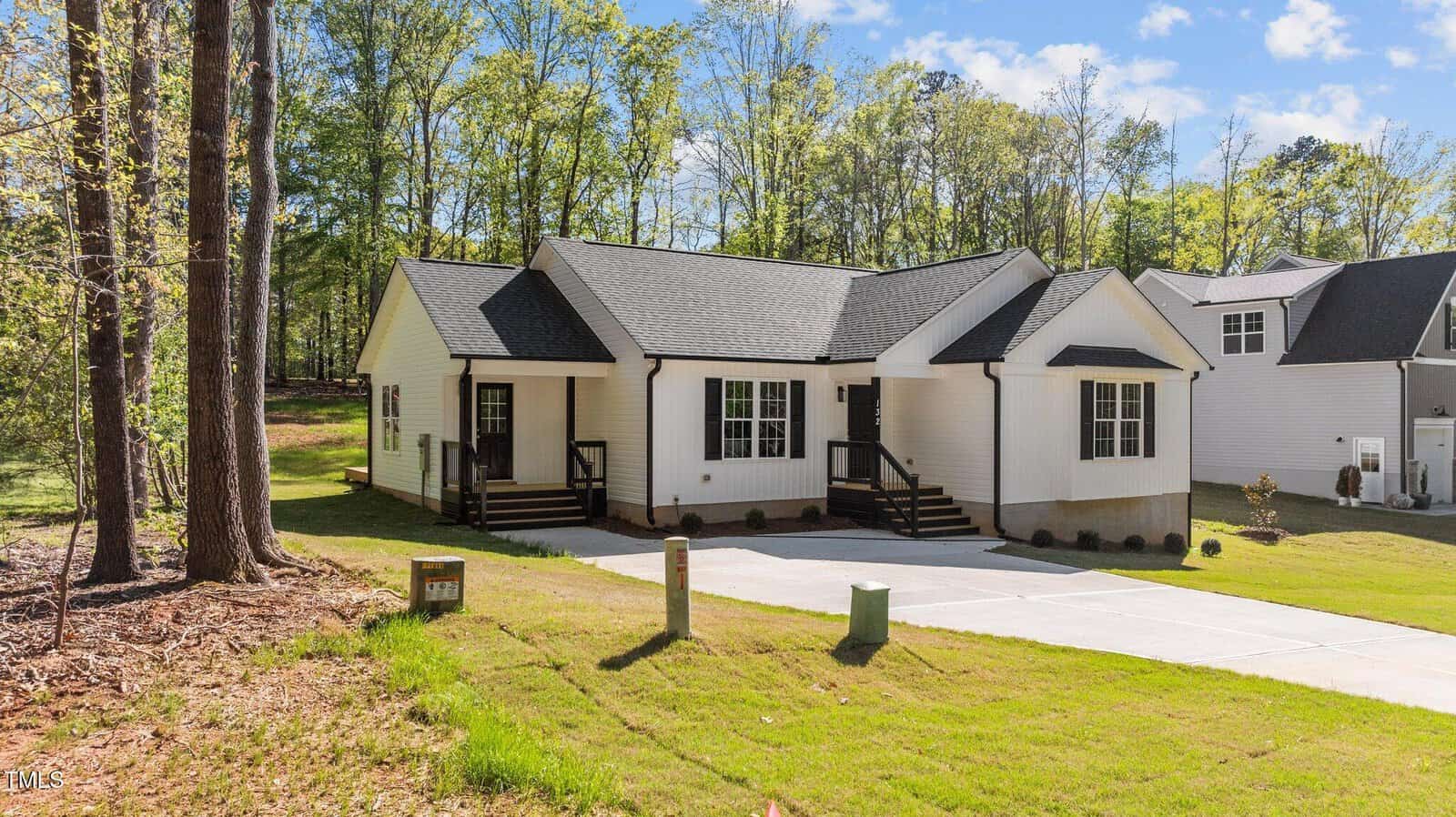 Single-story white house with black trim, a gabled roof, and two front entrances. The house sits on a grassy lot with a concrete driveway and wooded area in the background.