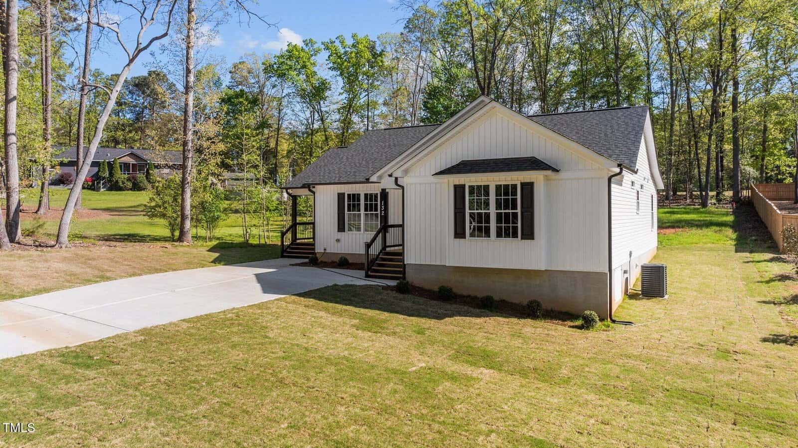 A single-story white house with black roof, front porch, and steps leading to the entrance sits on a large grassy yard with a concrete driveway. Tall trees and neighboring houses are visible in the background.