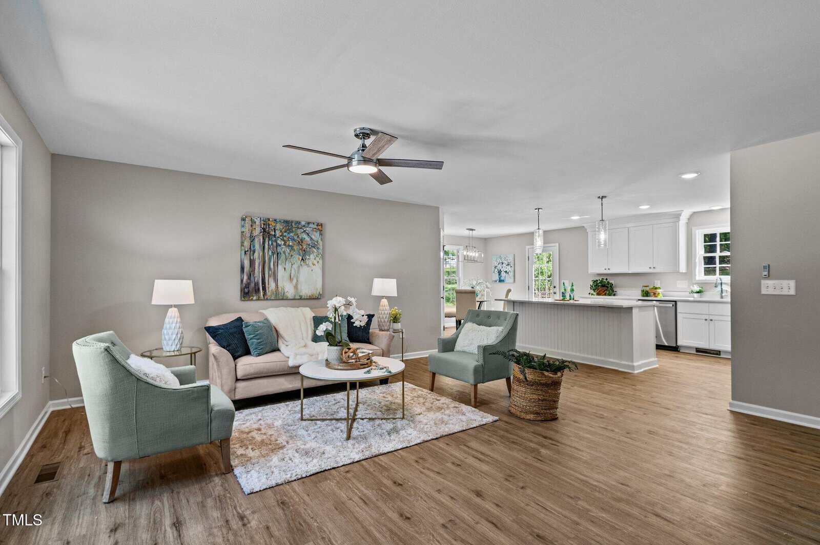 A modern open-concept living room with light-colored walls, wood floors, a beige sofa, two armchairs, a coffee table, and lamps. The kitchen with white cabinets and countertops is visible in the background.