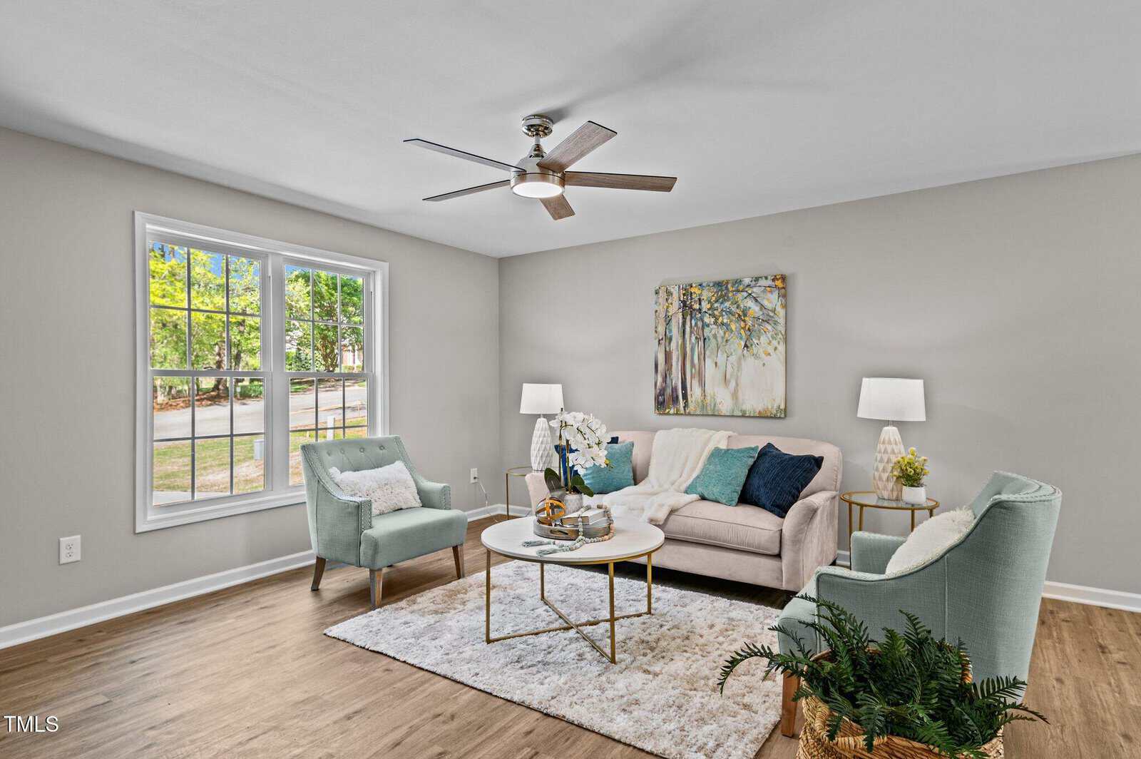 A living room with light wood flooring, a beige sofa, two light blue armchairs, a white rug, round coffee table, two side tables with lamps, a large window, a ceiling fan, and wall art above the couch.