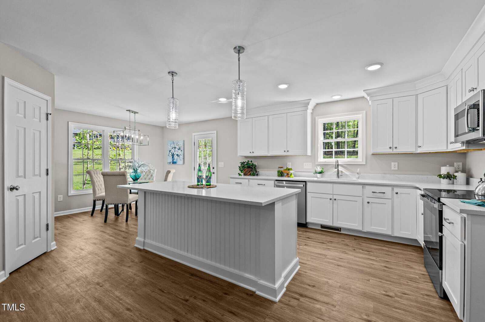 A modern kitchen with white cabinets, an island, stainless steel appliances, wood flooring, and pendant lights. Large windows let in natural light. A dining area with a table and chairs is visible in the background.