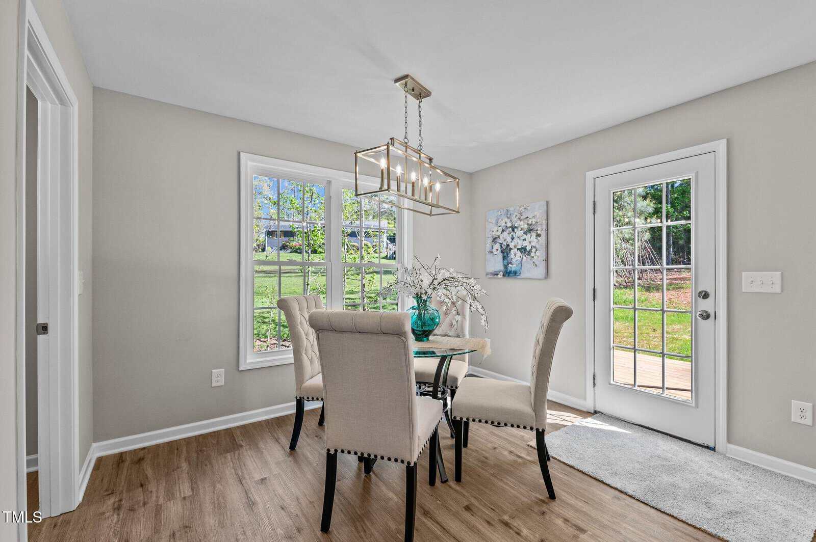 Bright dining area with a glass table, four upholstered chairs, a decorative vase, large windows showing a green yard, a modern light fixture, and a glass door leading outside.