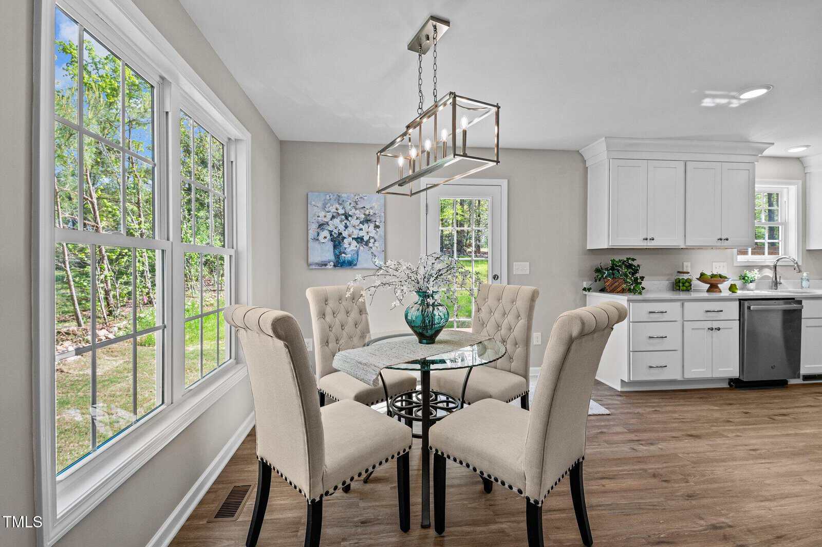 Bright kitchen with large windows, white cabinets, wood floor, and a small round glass dining table with four upholstered chairs, featuring a vase with white flowers as a centerpiece.