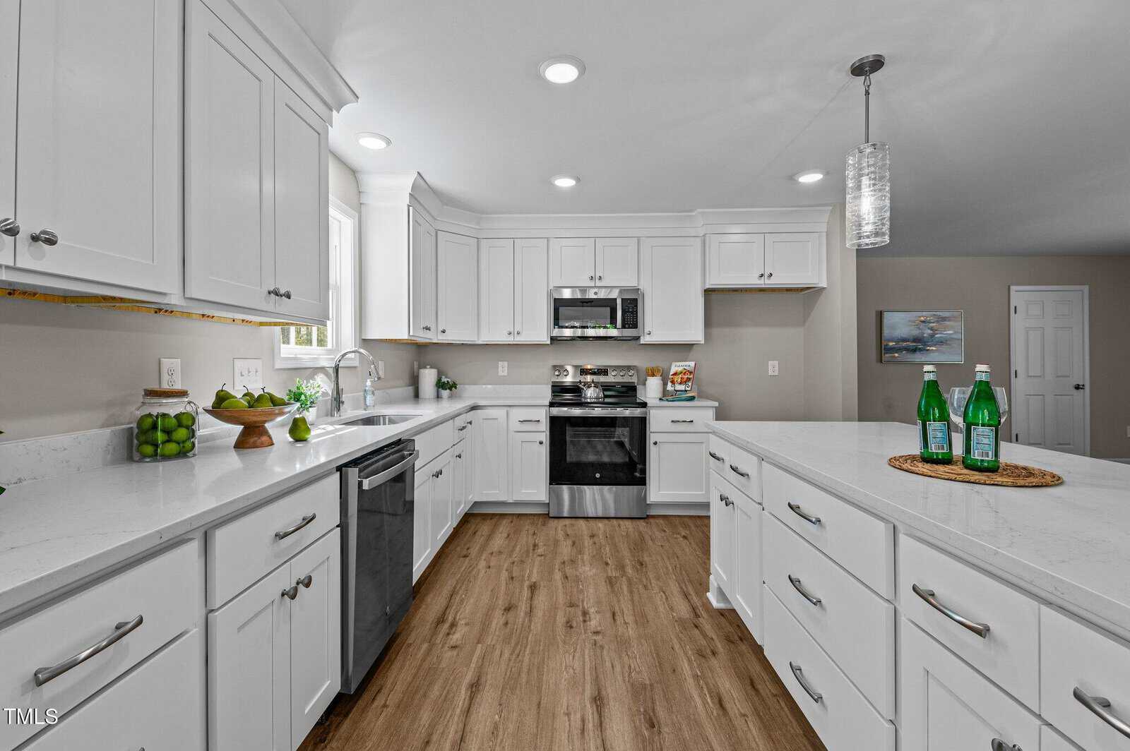 Modern white kitchen with stainless steel appliances, white cabinets, quartz countertops, wood flooring, and a center island topped with green glass bottles. Bright, clean, and open layout with recessed and pendant lighting.