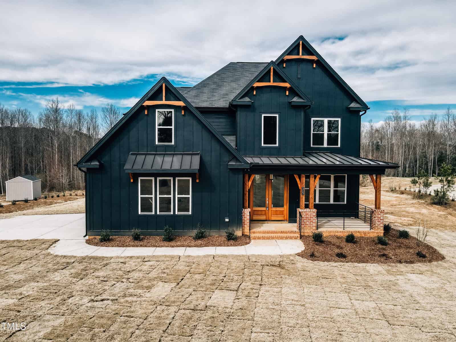 A modern two-story house with dark exterior siding, wood accents, a covered front porch, and large windows, situated on a spacious yard with minimal landscaping and surrounded by trees.