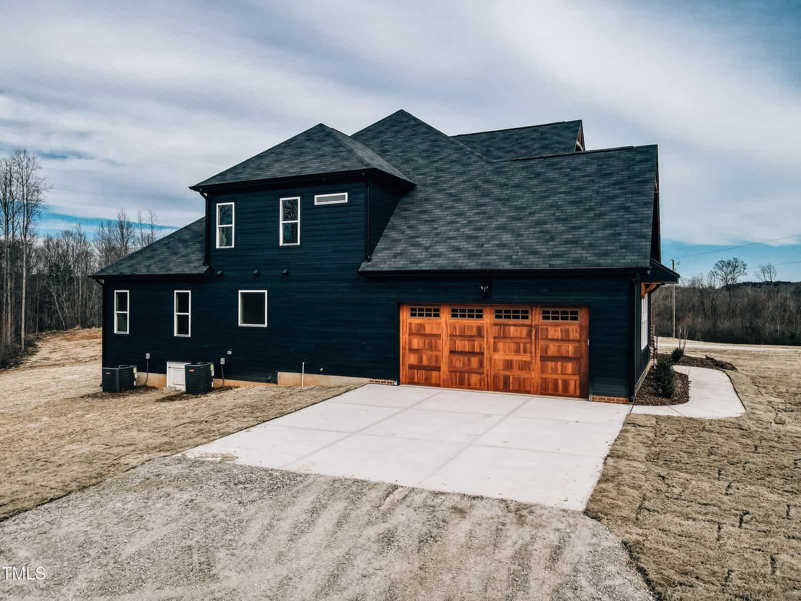 A modern two-story house with dark siding and a wooden double garage door sits on a spacious lot with a concrete driveway and minimal landscaping. The sky is mostly cloudy, and trees are visible in the background.