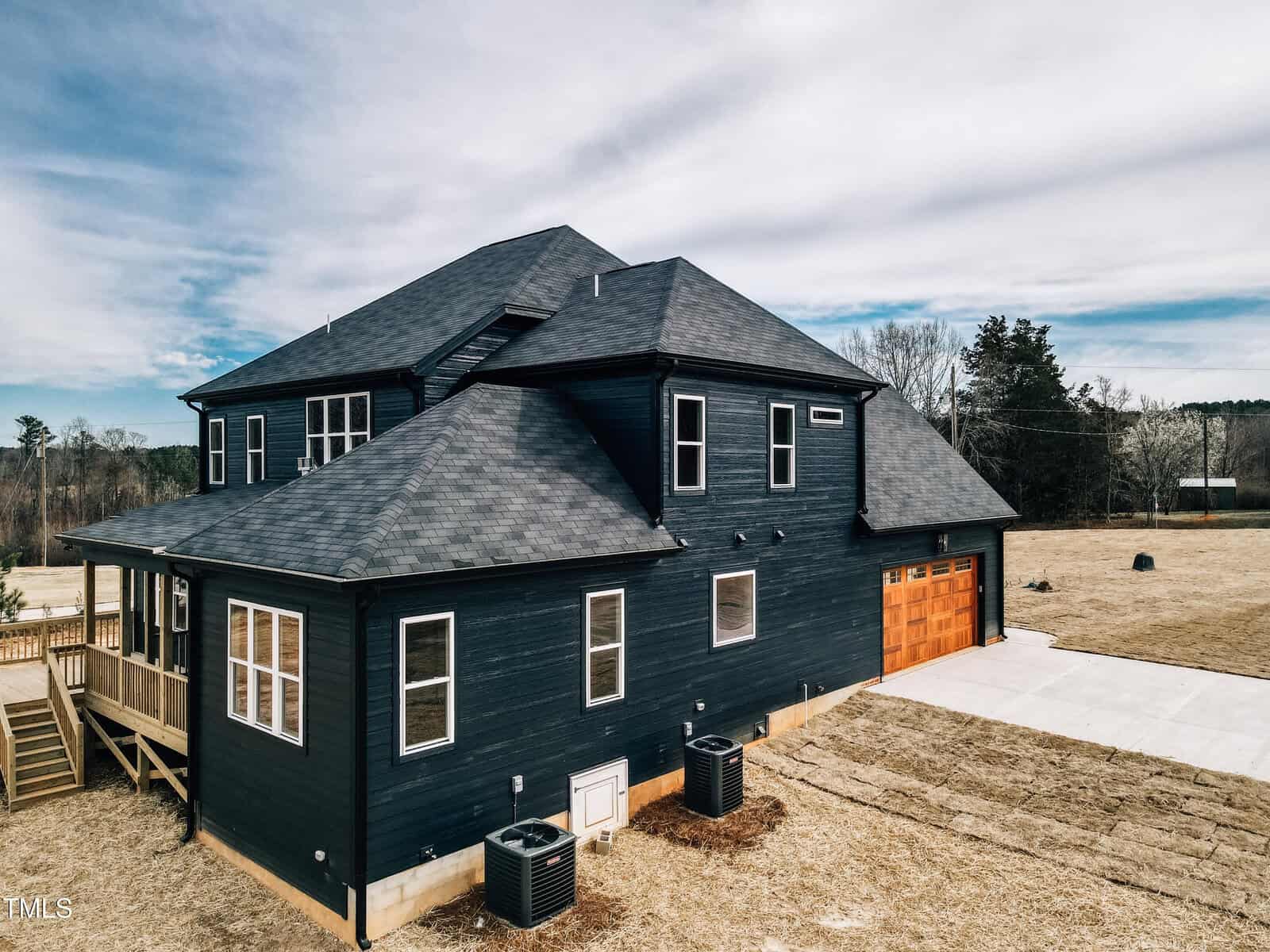 A two-story dark blue house with a gray shingle roof, wooden garage doors, and multiple white-trimmed windows. The yard is mostly bare with some grass and trees in the background. Two HVAC units are beside the house.
