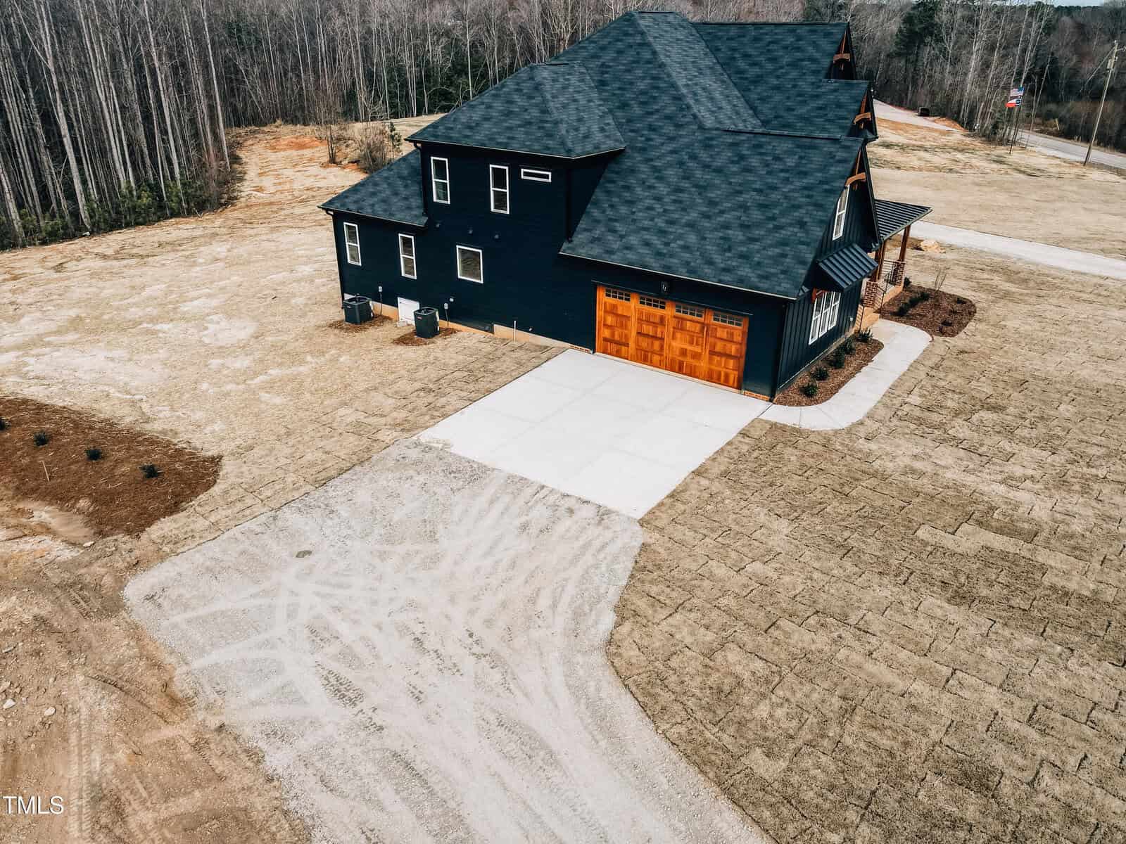 A modern black house with a wooden garage door sits on a large, mostly bare lot with a paved driveway and sparse patches of newly laid sod. The surrounding area is partially wooded.