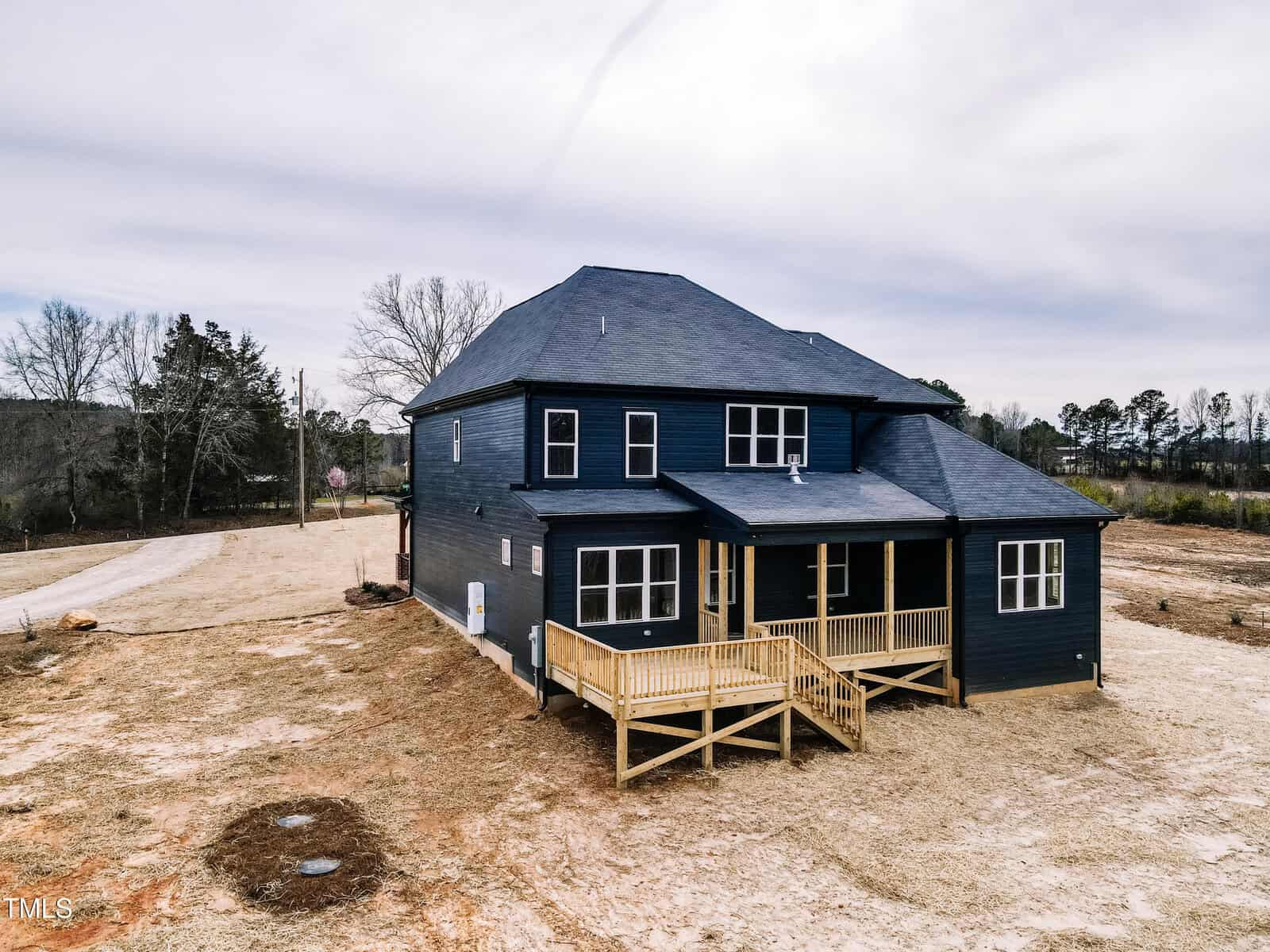 A two-story dark blue house with a covered porch and wooden deck sits on a large, mostly bare yard with trees and a road in the background under a cloudy sky.