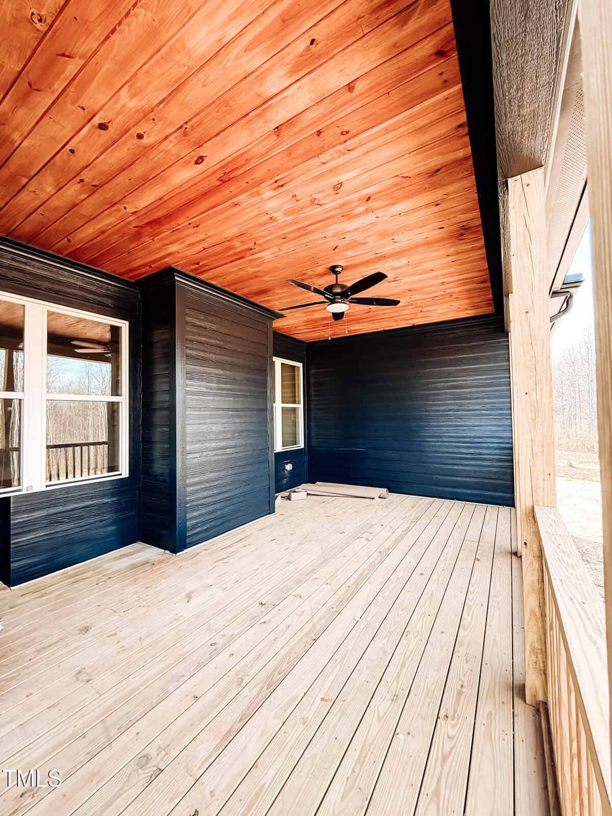 A spacious covered porch with light wooden floors and ceiling, dark painted walls, windows, a ceiling fan, and open view to the outdoors.