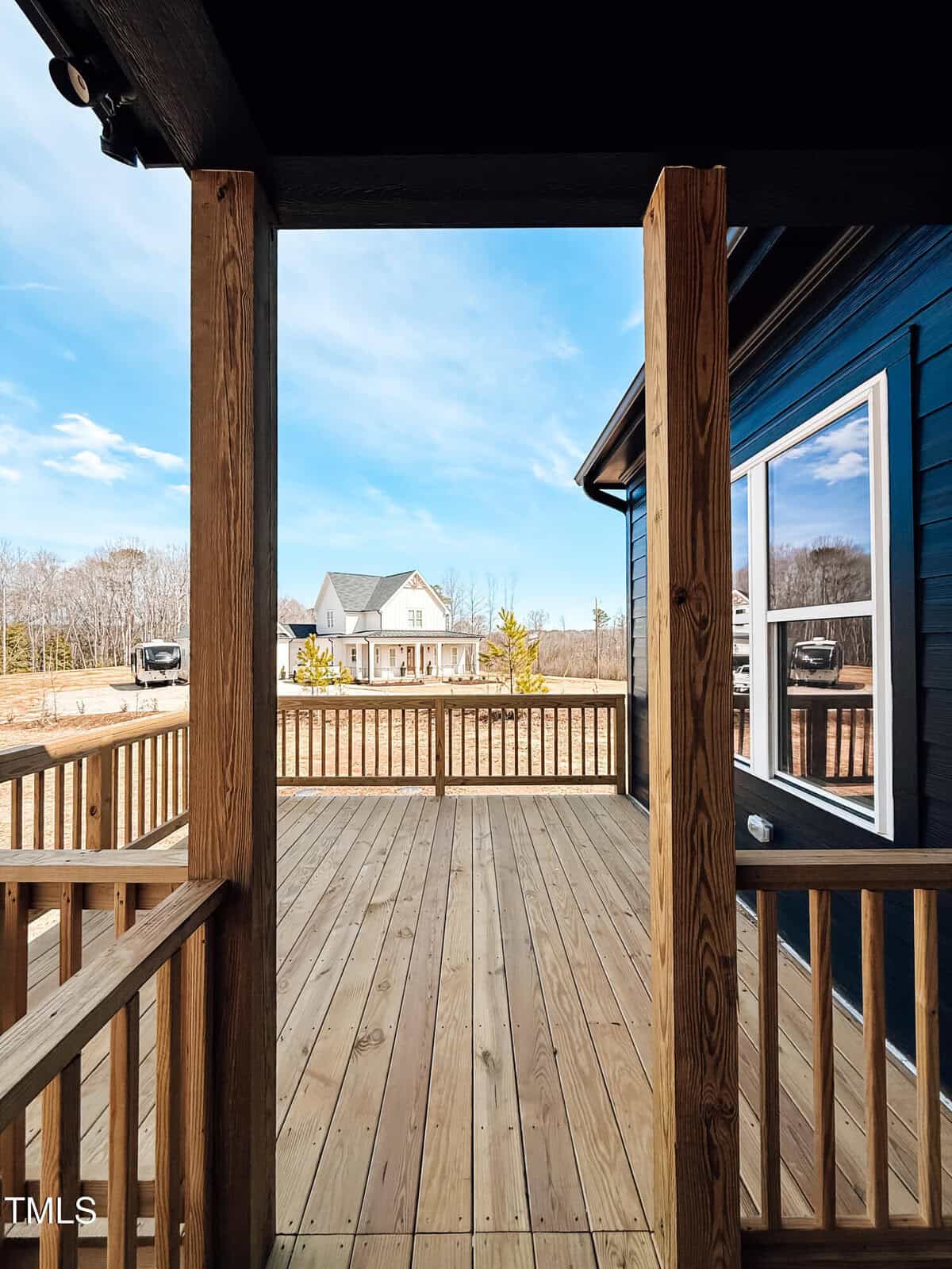 View from a wooden porch looking out to a spacious yard with a white house in the distance, under a blue sky with scattered clouds. The porch has wooden railings and posts.