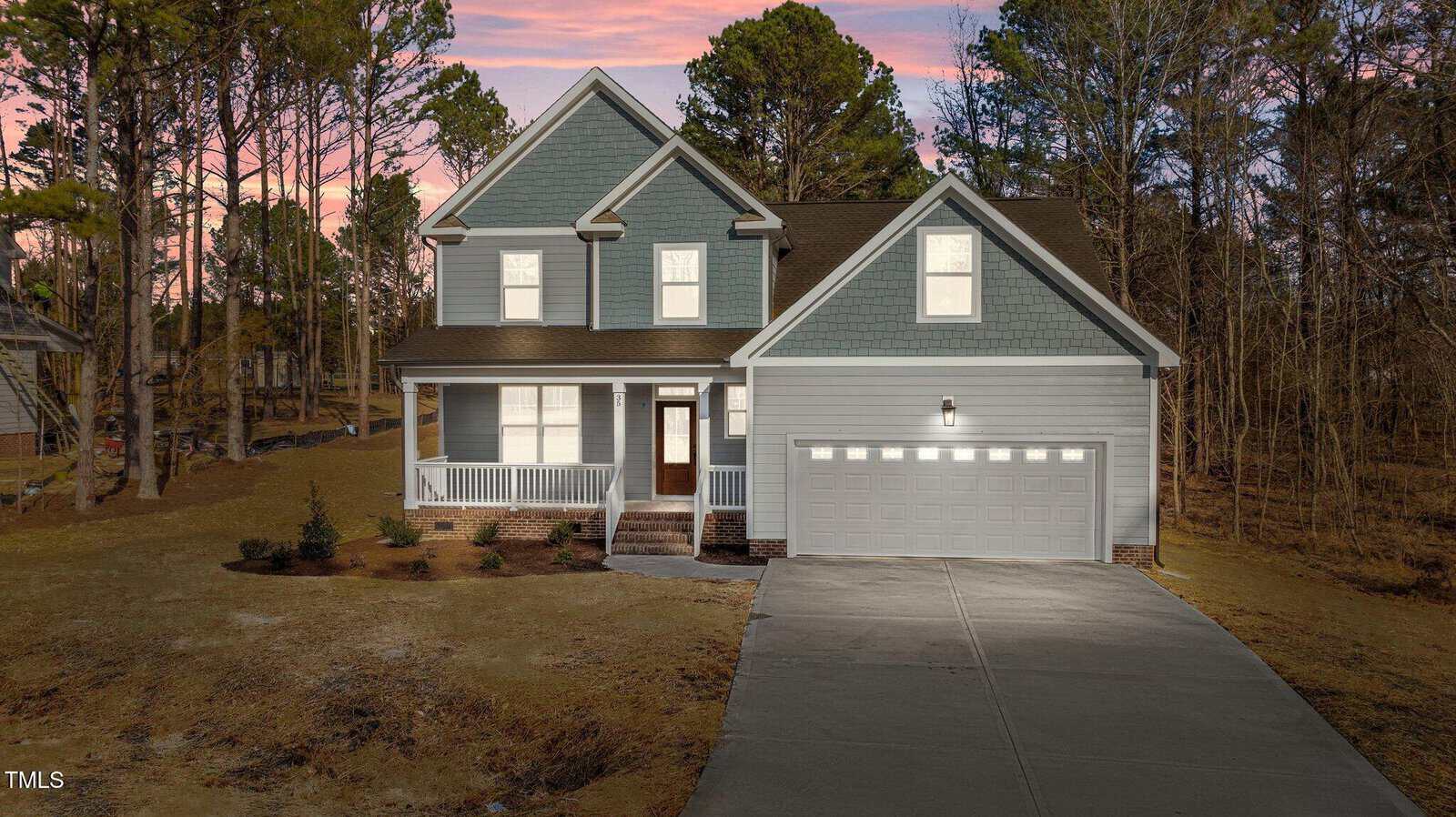 A two-story blue house with white trim, a front porch, and a two-car garage. The driveway is concrete, and the yard is mostly bare soil with a few small shrubs. Trees surround the property at sunset.