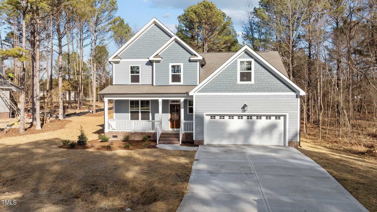 A two-story gray house with white trim, a covered front porch, and attached two-car garage sits on a large lot with a concrete driveway and surrounding pine trees. The yard has minimal landscaping.