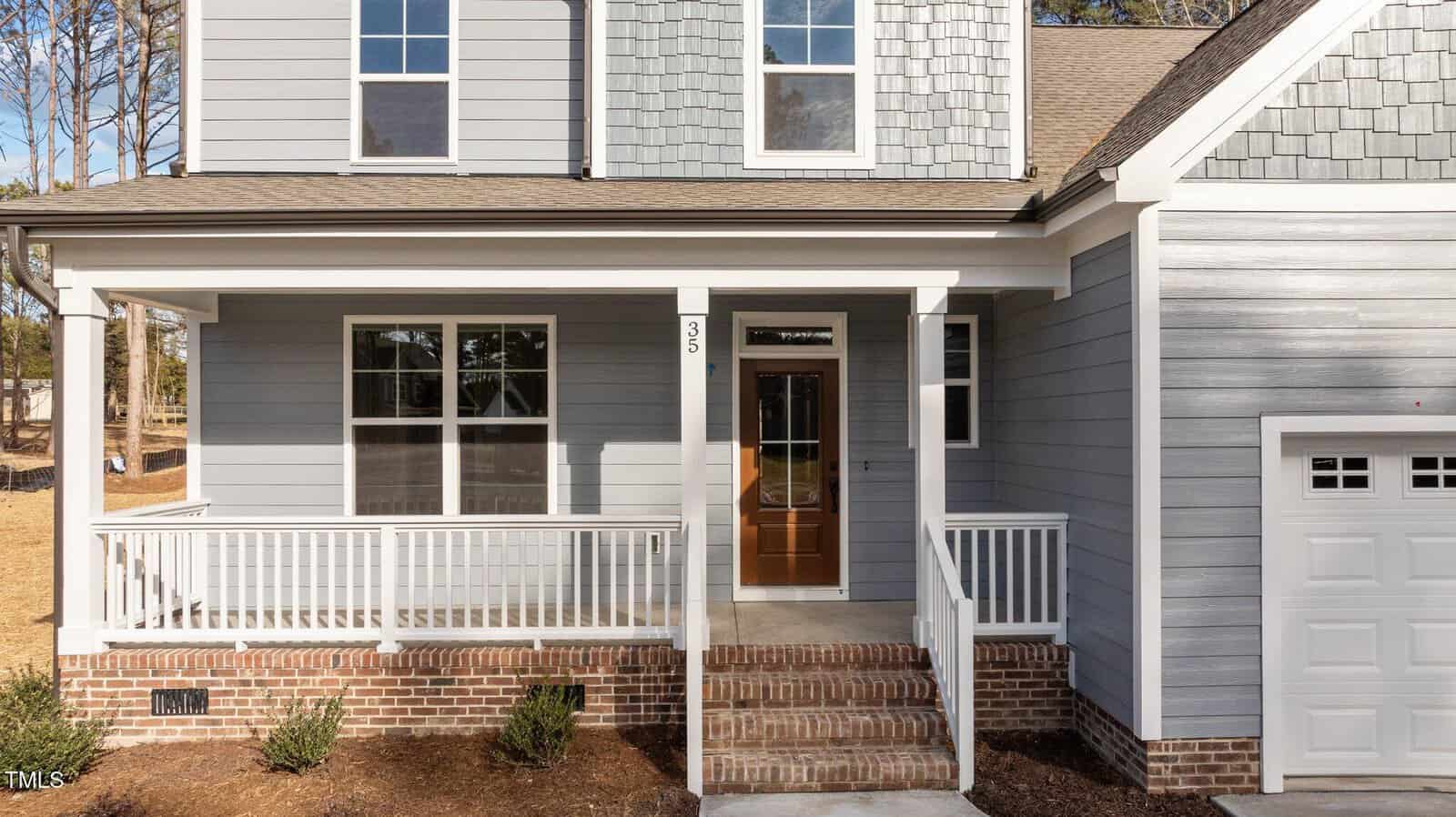 A front view of a modern house with light gray siding, brick foundation, a covered porch with white railings, a wooden front door with glass panels, and a garage partially visible on the right.