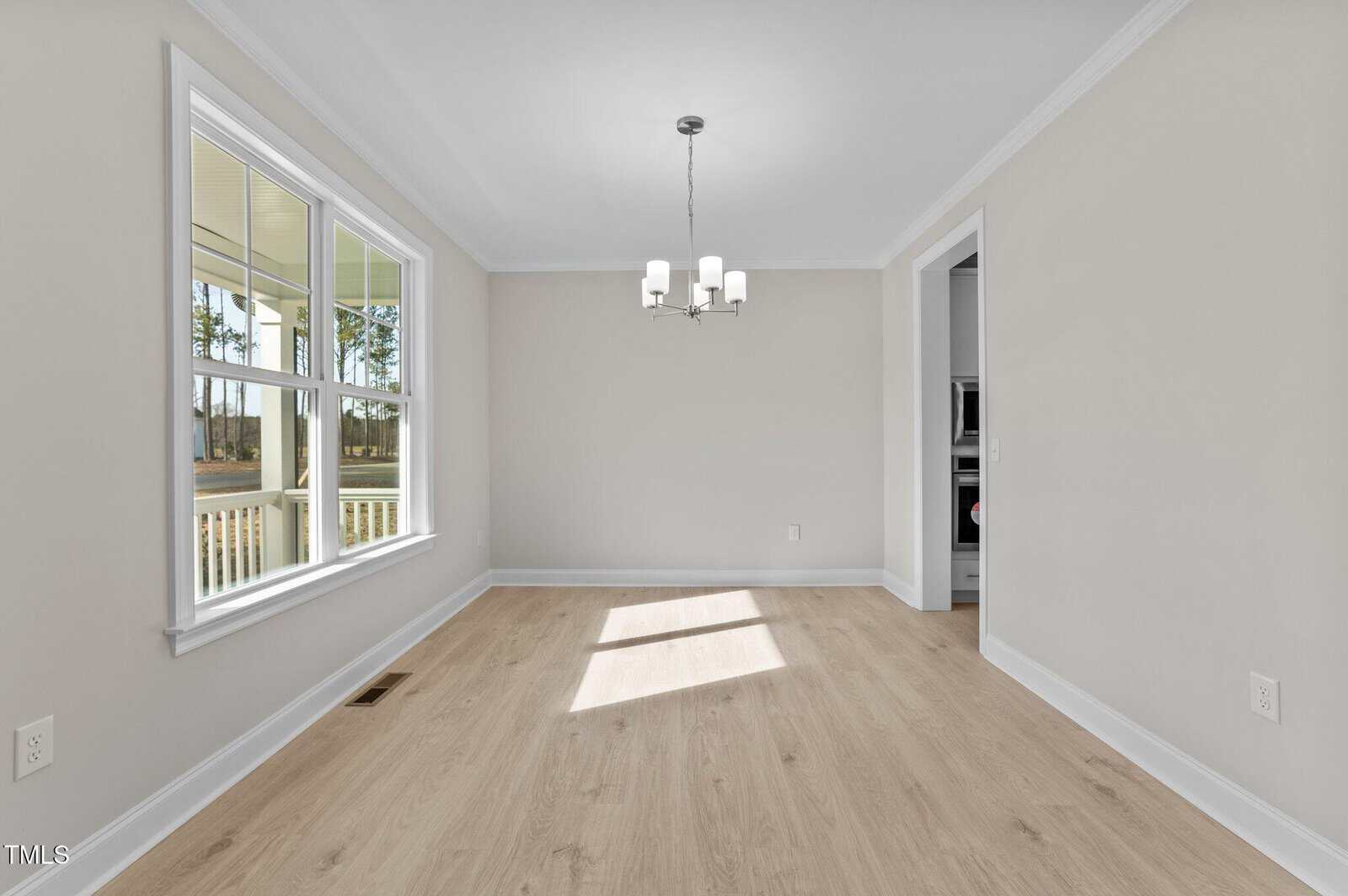 Bright, empty dining room with light wood floors, beige walls, a large window, white trim, and a modern chandelier. Natural light fills the space; adjacent to a kitchen entrance.