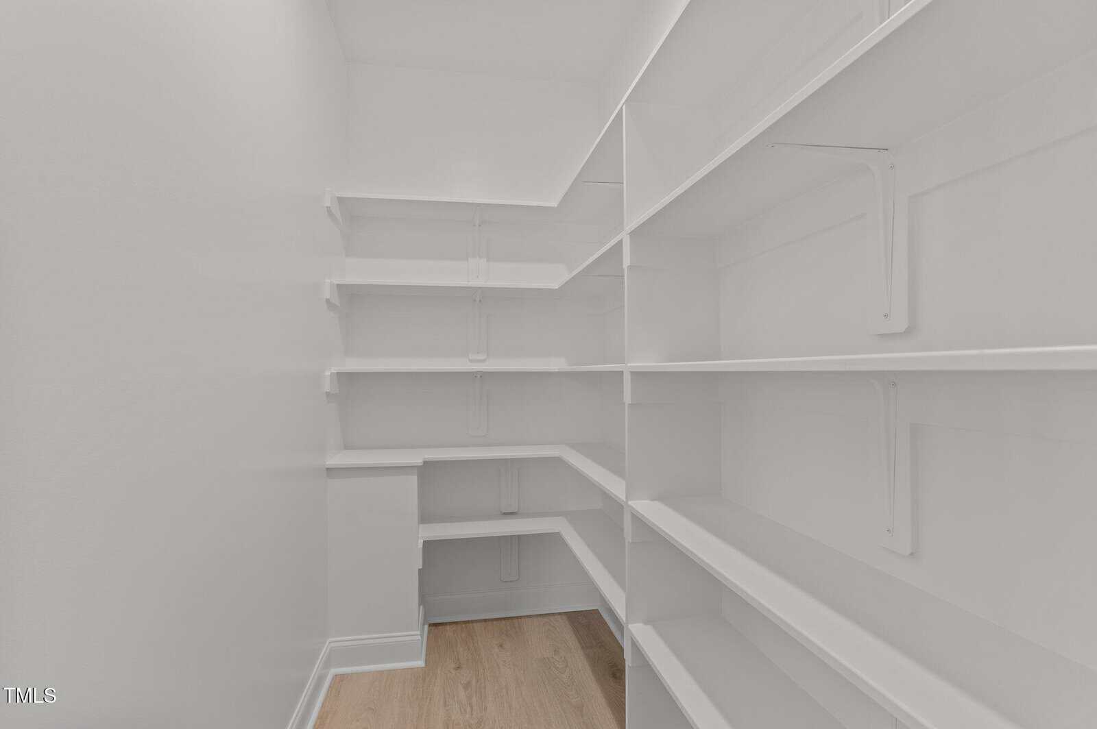 A walk-in pantry with white shelves lining the walls on both sides and light wood flooring. The space is empty and well-lit, with multiple levels of shelving for storage.