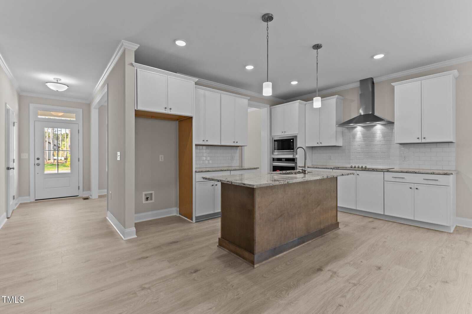 Modern kitchen with white cabinets, stainless steel appliances, a tile backsplash, and a center island with a sink. Pendant lights hang above the island. Light wood flooring and neutral walls throughout the space.