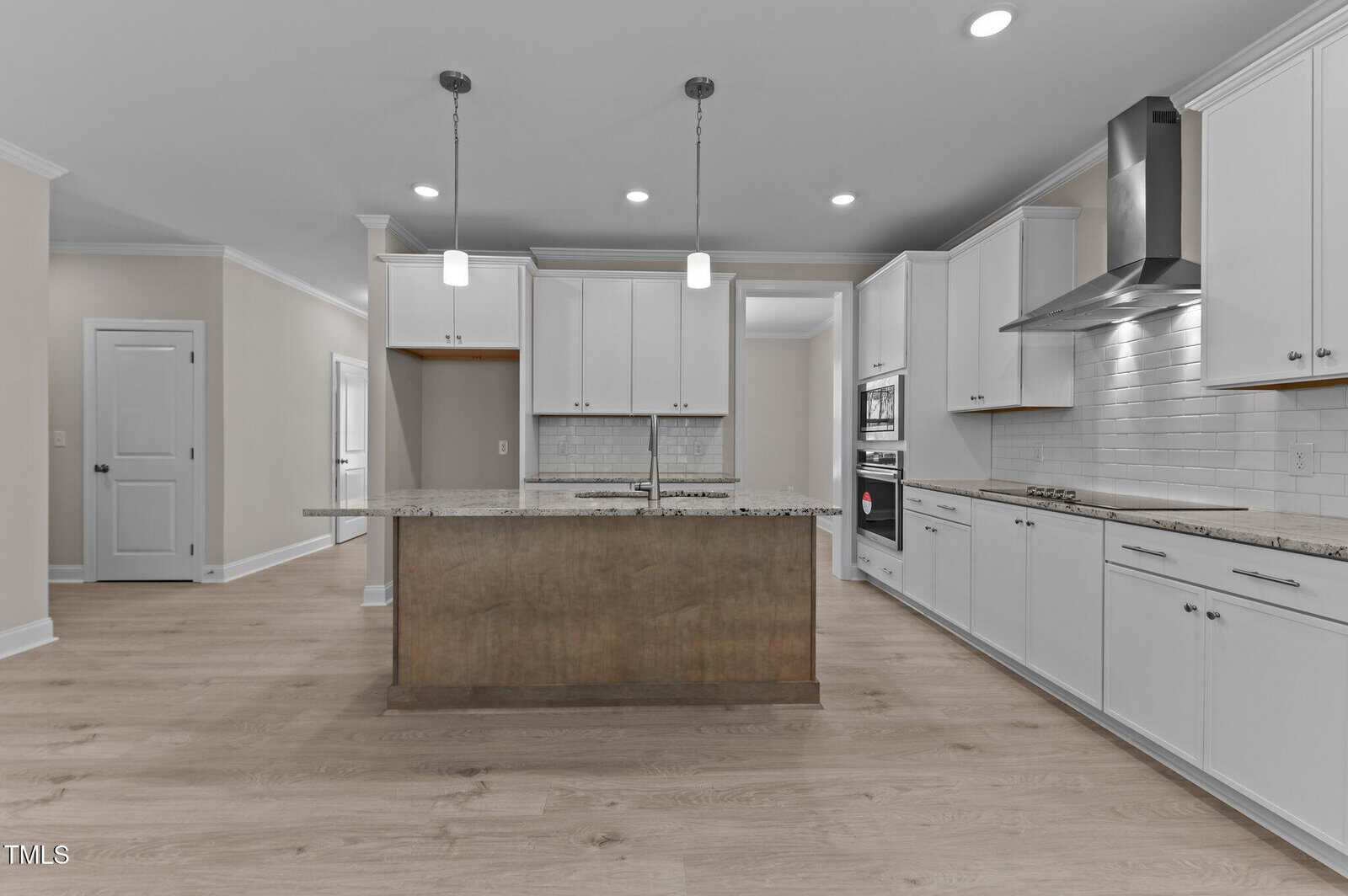 A modern kitchen with white cabinets, a stainless steel range hood, built-in oven, subway tile backsplash, granite island with sink, and light wood flooring under recessed and pendant lighting.