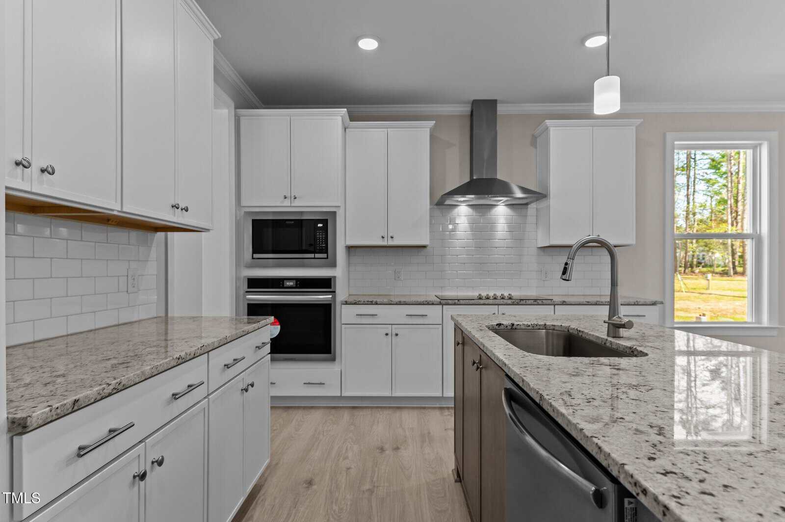 Modern kitchen with white cabinets, granite countertops, built-in microwave, stainless steel appliances, island with sink, under-cabinet lighting, and a window showing trees outside. Light wood flooring throughout.
