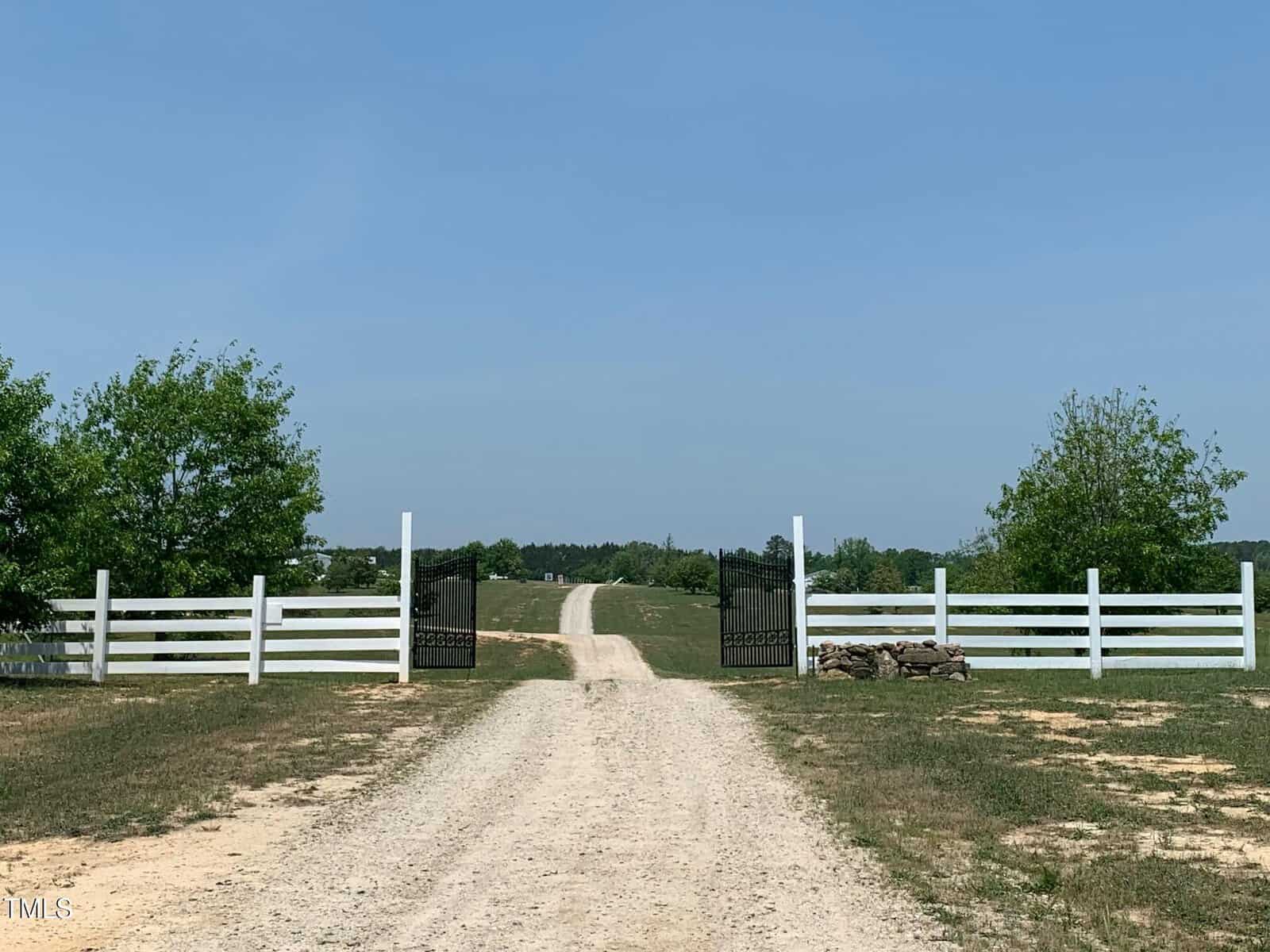 A dirt road leads through an open gate with a white fence on both sides at 1800 Gillburg Road. The path stretches into the distance under a clear blue sky, flanked by green trees and grass. Rocks are piled near the gate, painting a serene picture of Henderson's countryside.