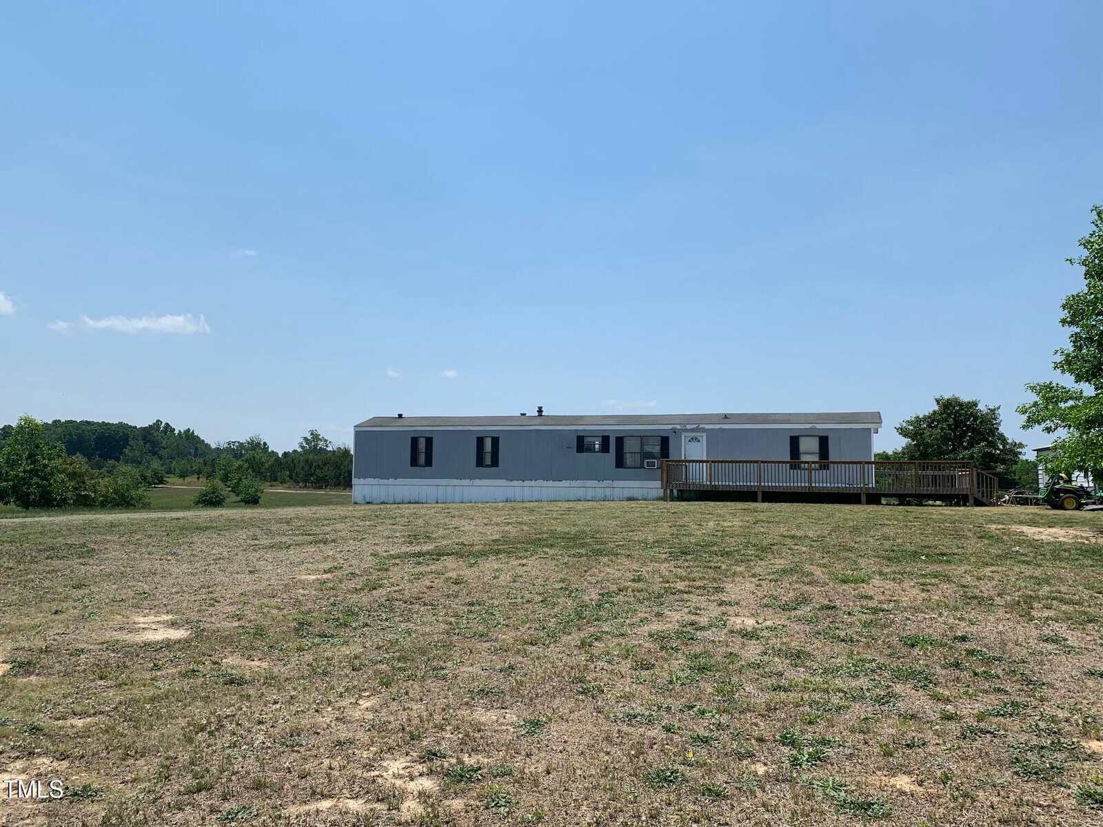 A single-story mobile home sits on a vast, grassy field under a clear blue sky. The white structure has a wooden ramp leading to the entrance. Sparse greenery and trees are visible in the distance.