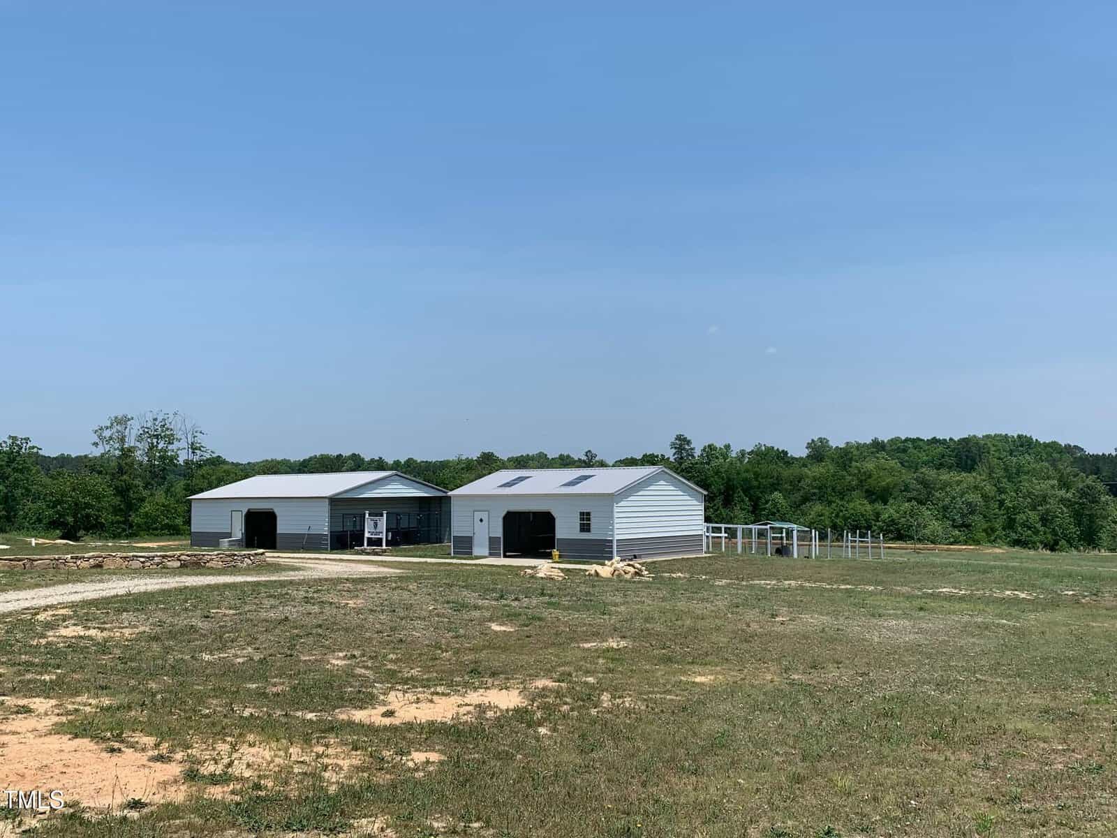 A rural landscape with two white barn-like structures surrounded by green grass and trees under a clear blue sky. A dirt path leads to the buildings, and there's a fenced area beside them.