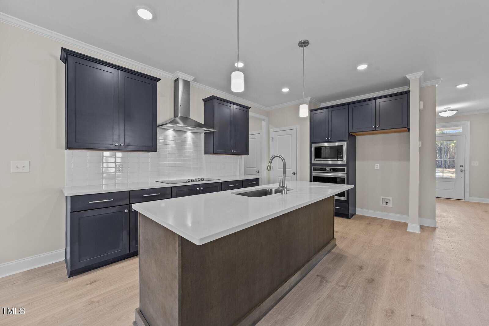 Modern kitchen with dark cabinets, white countertops, a center island with sink, built-in oven and microwave, stainless steel hood, white subway tile backsplash, pendant lights, and light wood flooring.