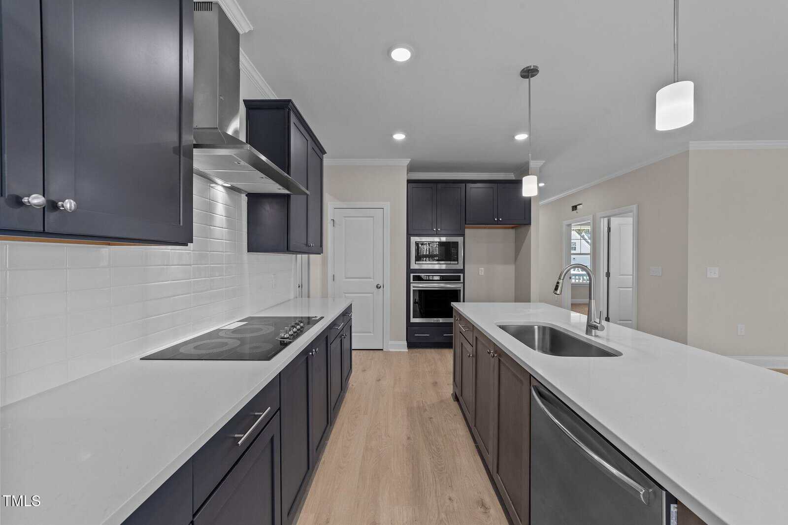 Modern kitchen with dark cabinets, white countertops, a cooktop, built-in oven and microwave, stainless steel sink, pendant lights, and light wood flooring. White subway tile backsplash is behind the stove area.