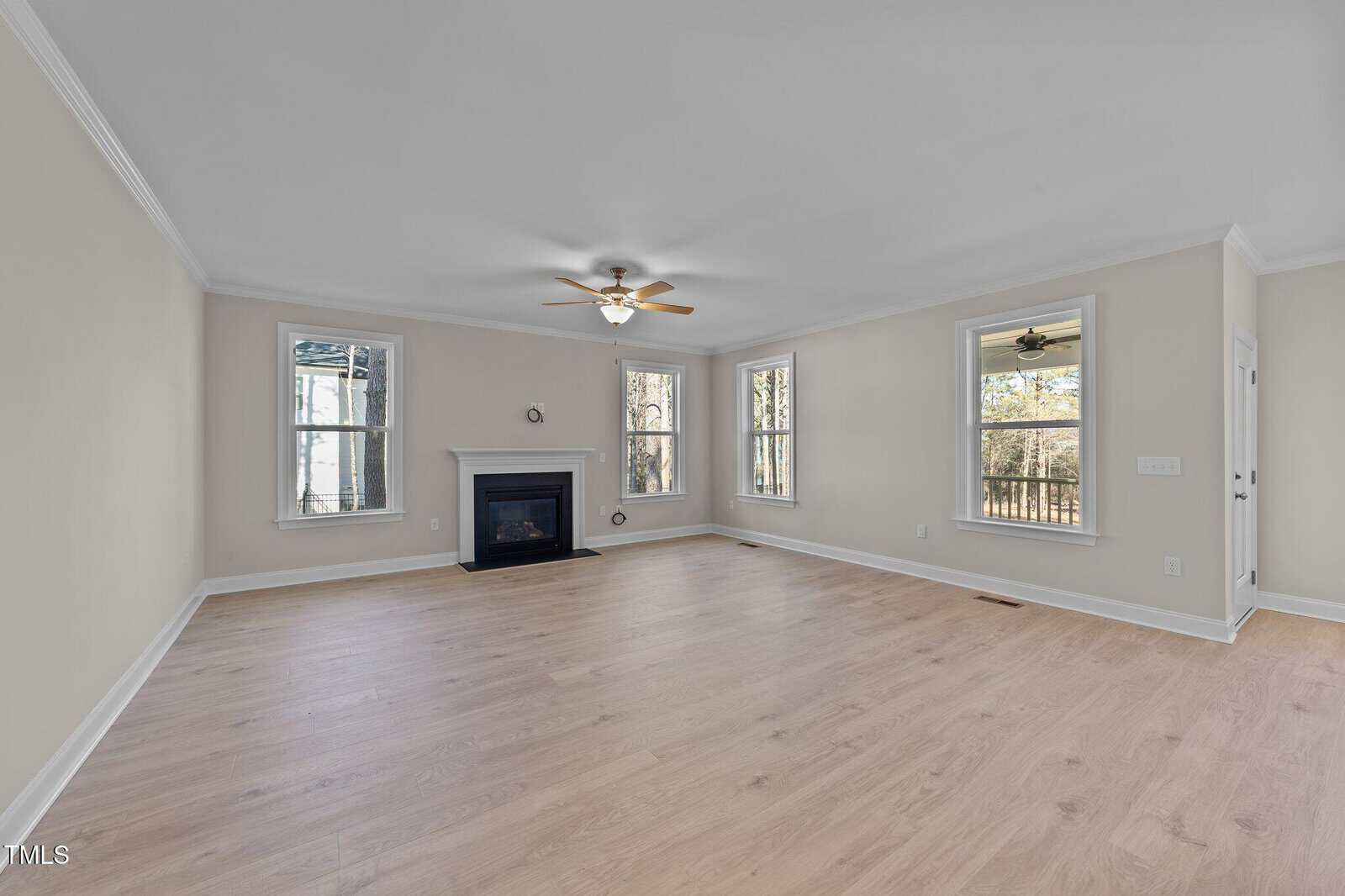 Spacious empty living room with light wood floors, beige walls, a ceiling fan with light, three large windows, a white-trimmed fireplace, and a door leading to a screened porch. Natural light fills the space.
