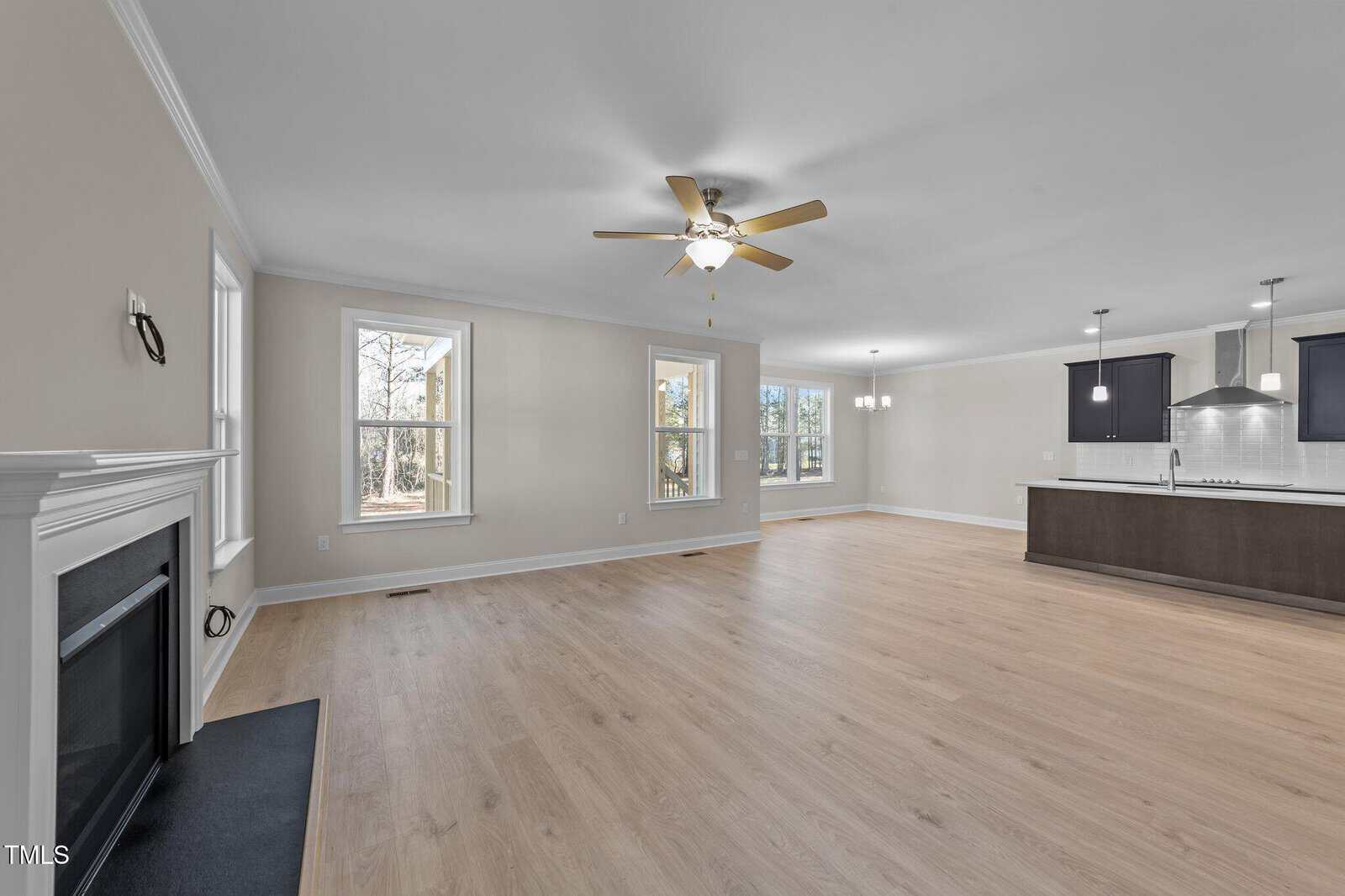 Spacious open-concept living area with light wood flooring, a ceiling fan, multiple large windows, a fireplace on the left, and a modern kitchen with dark cabinets and hanging lights in the background.