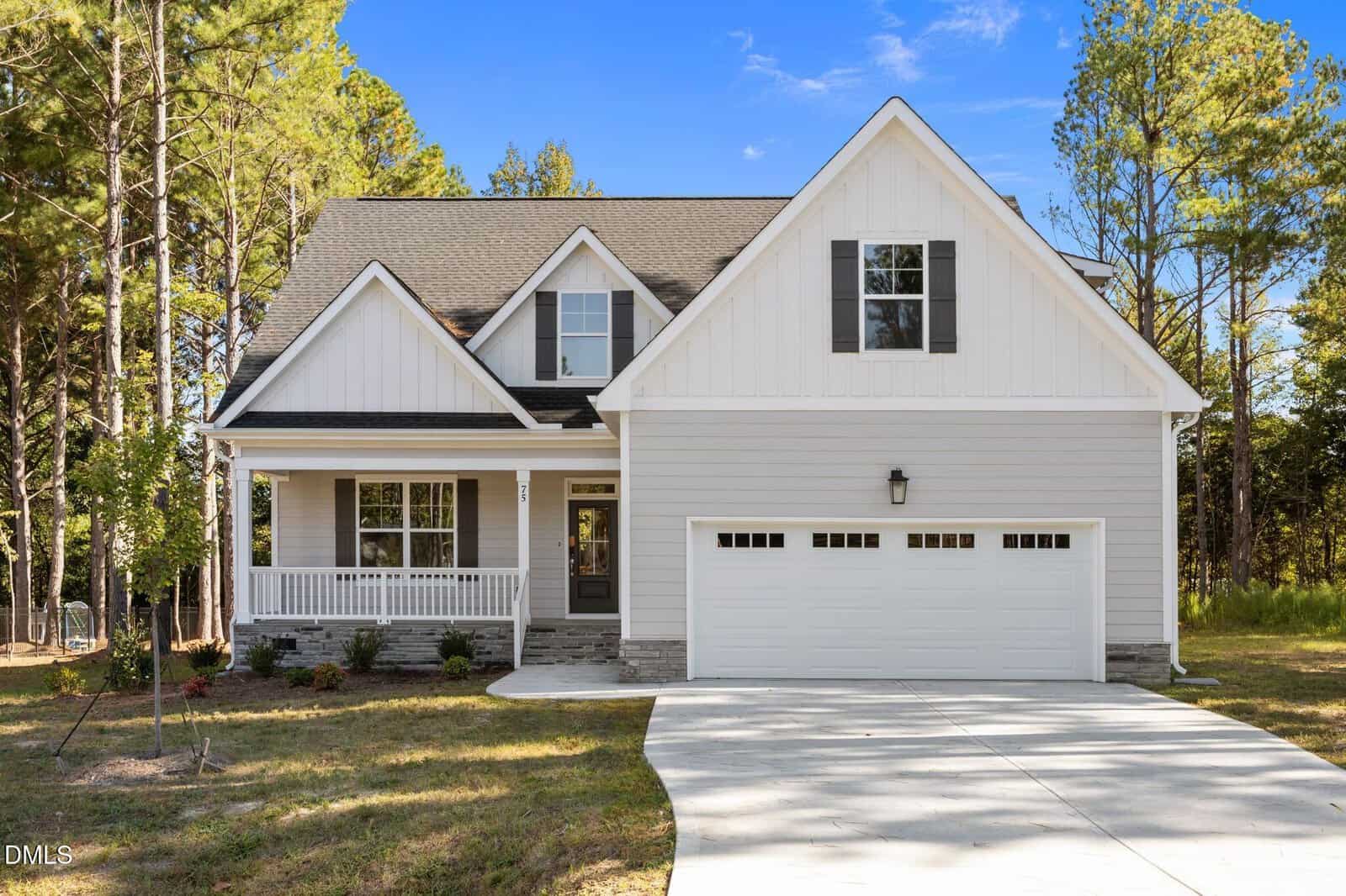 Nestled among tall pine trees at 75 Arbor Drive in Youngsville, this two-story house features light gray siding, white trim, a covered front porch, and a double garage. A concrete driveway leads to the garage, and the yard offers minimal landscaping for easy maintenance.