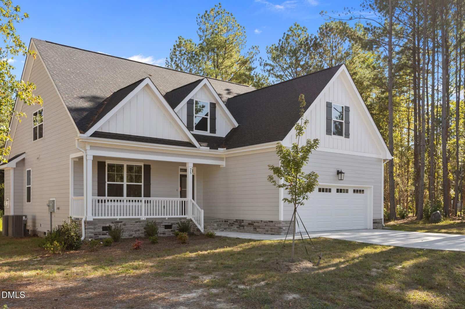 A white two-story house with a covered front porch, two dormer windows, and a double garage. The house is surrounded by trees, and there is a small tree and minimal landscaping in the front yard.