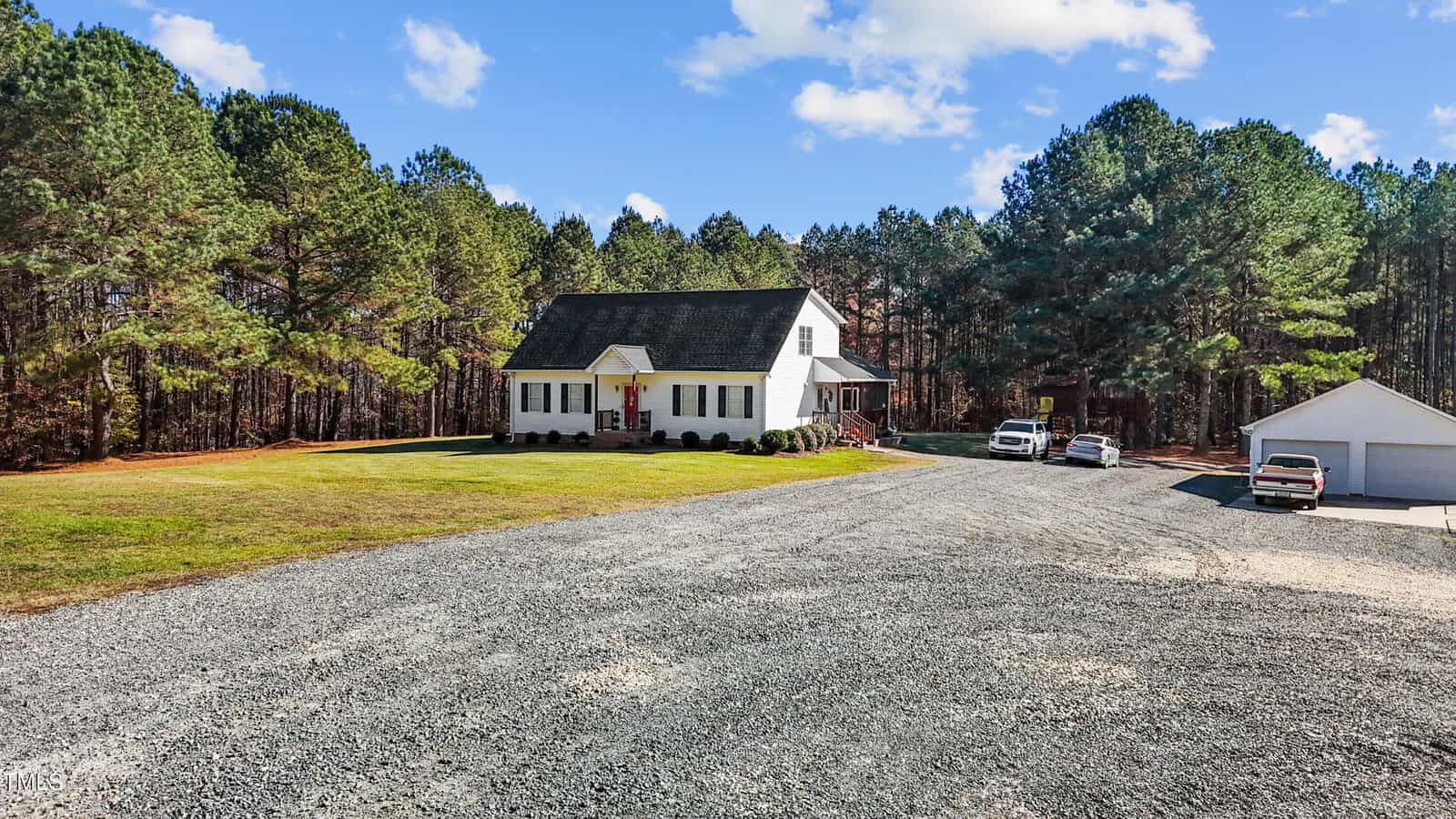 Situated at 3214 Tump Wilkins Road, this white house with a gabled roof sits on a spacious lot surrounded by trees. A large gravel driveway leads to the house, where two vehicles are parked alongside a detached garage. The sky is partly cloudy, adding to the serene atmosphere of this home.