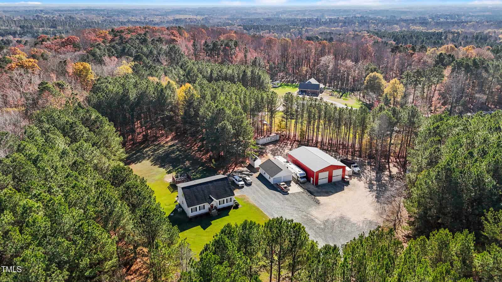 Aerial view of a wooded area featuring a house, a large red-and-white barn, and several vehicles parked on a gravel driveway. The property is surrounded by dense forest, with another house visible in the distance.