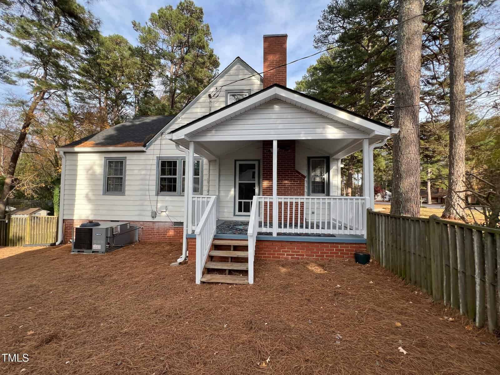 A small, white house with a brick chimney and a covered back porch with steps leading down. The yard is covered in pine needles, bordered by a wooden fence, and surrounded by tall trees. An air conditioning unit is visible on the side.