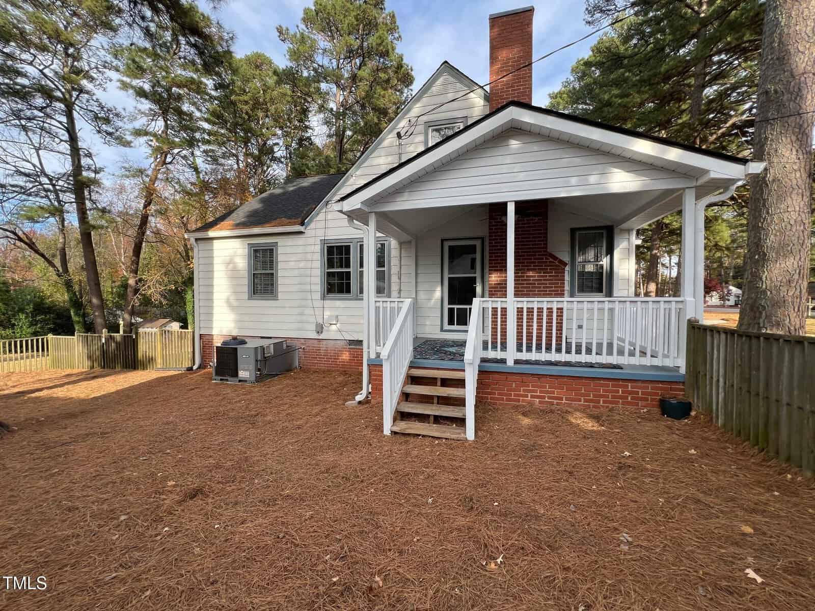 A white house with a brick foundation and a covered porch surrounded by tall pine trees. The yard is covered in pine straw, with a wooden fence enclosing the space. An HVAC unit is visible on the left side of the house.