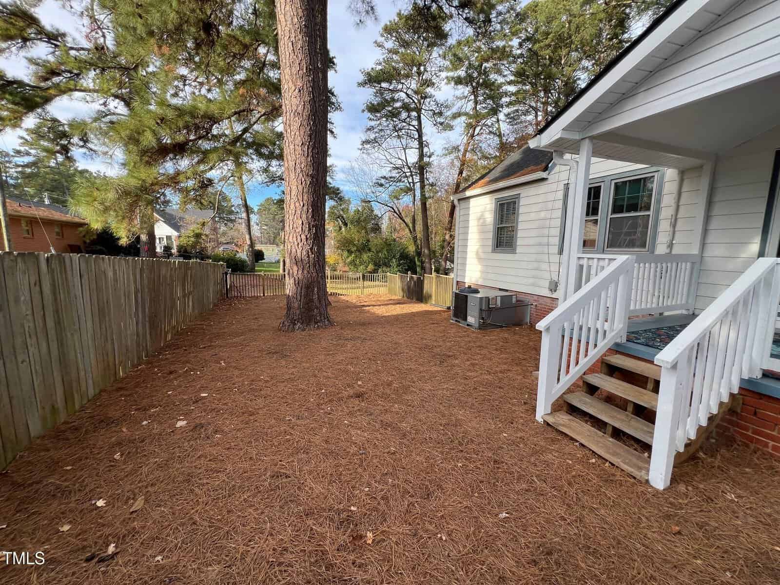 A backyard with a white house and wooden steps, surrounded by a tall wooden fence and large pine trees. The ground is covered with pine needles. An air conditioning unit is visible next to the house.