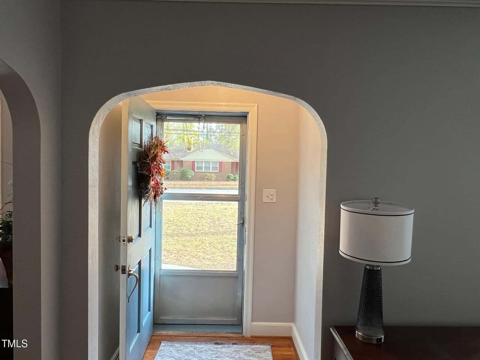 Open front door with a wreath, revealing a view of a suburban street with a red brick house. Inside, a lamp sits on a side table next to the doorway, which has an arched frame. Sunlight illuminates the interior.