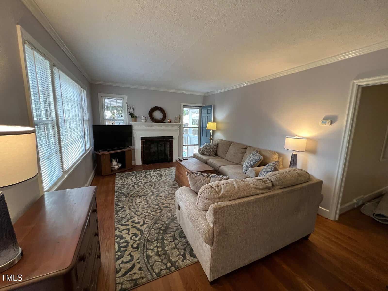 A cozy living room with a large beige sectional sofa and a patterned area rug. A fireplace with decorative items is along one wall, while large windows let in natural light. A wooden sideboard and a standing lamp flank the sofa.