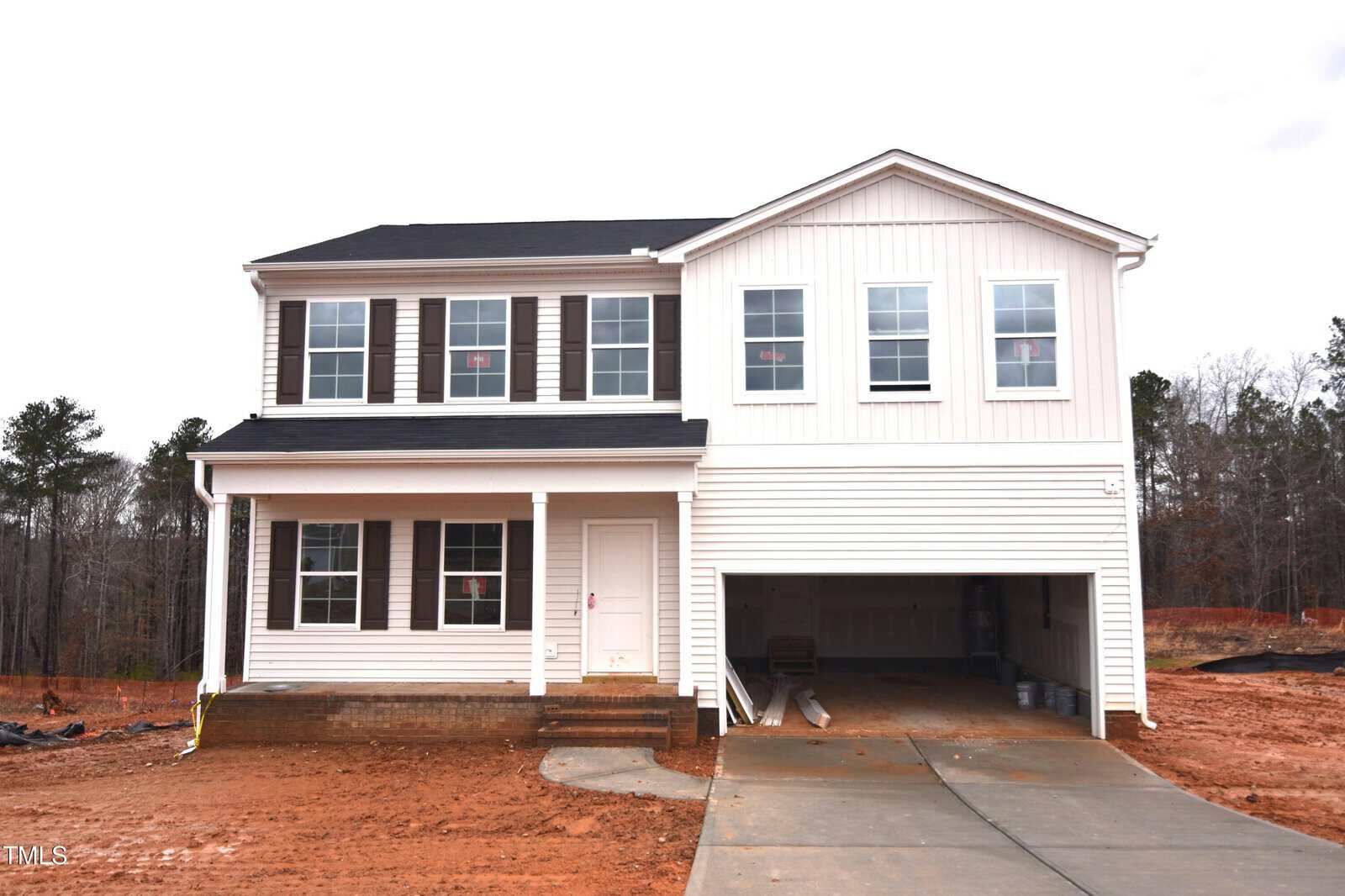 A two-story, white suburban house with a covered front porch, black shutters, and an attached two-car garage. The yard and driveway are unlandscaped with visible bare soil. Trees are in the background.