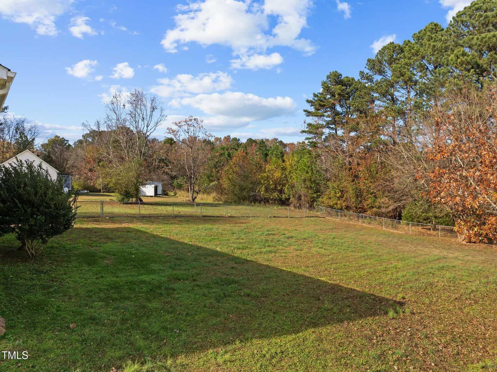 A backyard with a grassy lawn and a mix of trees in the background. The sky is partly cloudy, and a small white shed is visible on the left side near the tree line. The area is surrounded by a wire fence, with a house partially seen on the left.