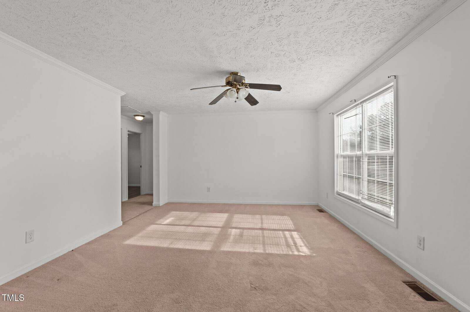 A minimalist living room with plain white walls and beige carpet. A ceiling fan with brown blades is centrally mounted. Sunlight streams through a window with blinds, casting shadows on the carpet. An open doorway leads to another room.