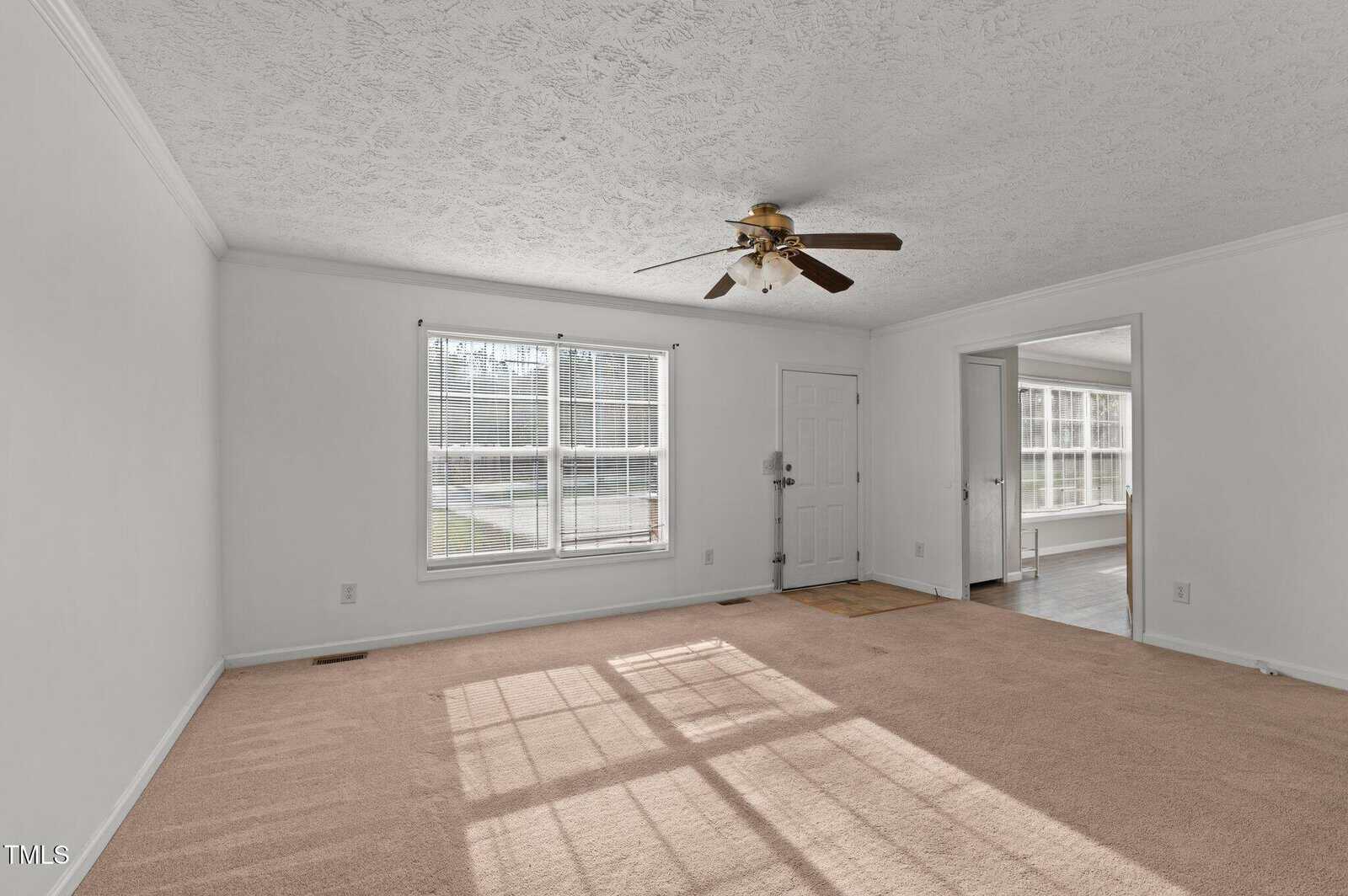 A bright, empty living room with a beige carpet and textured white ceiling. A ceiling fan is centered above. A large window and a door are on one wall. An open doorway leads to another room with additional windows and light wood flooring.