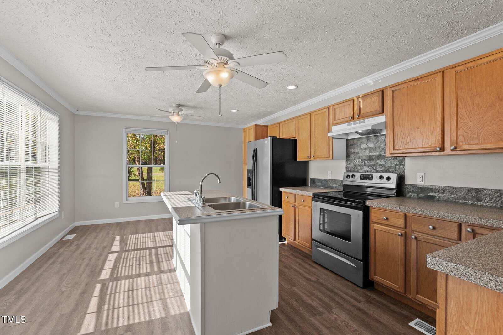 A modern kitchen with wooden cabinets, a central island with a sink, and stainless steel appliances, including a stove and refrigerator. There are two ceiling fans, large windows, and wood flooring.